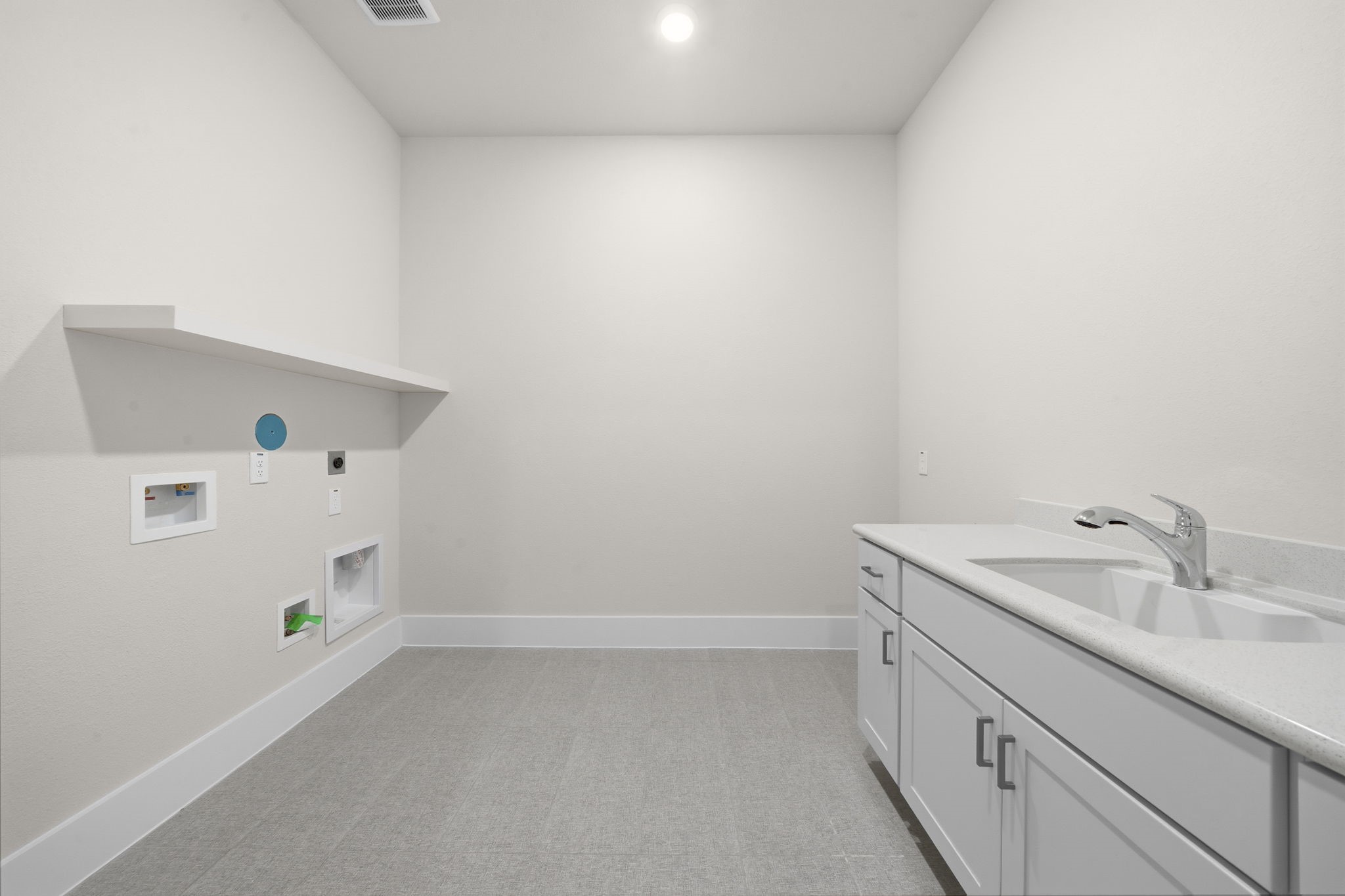 Spacious laundry room with light-colored walls and flooring, featuring a countertop with a sink and ample cabinetry for storage. A shelf and hookups are available for appliances.