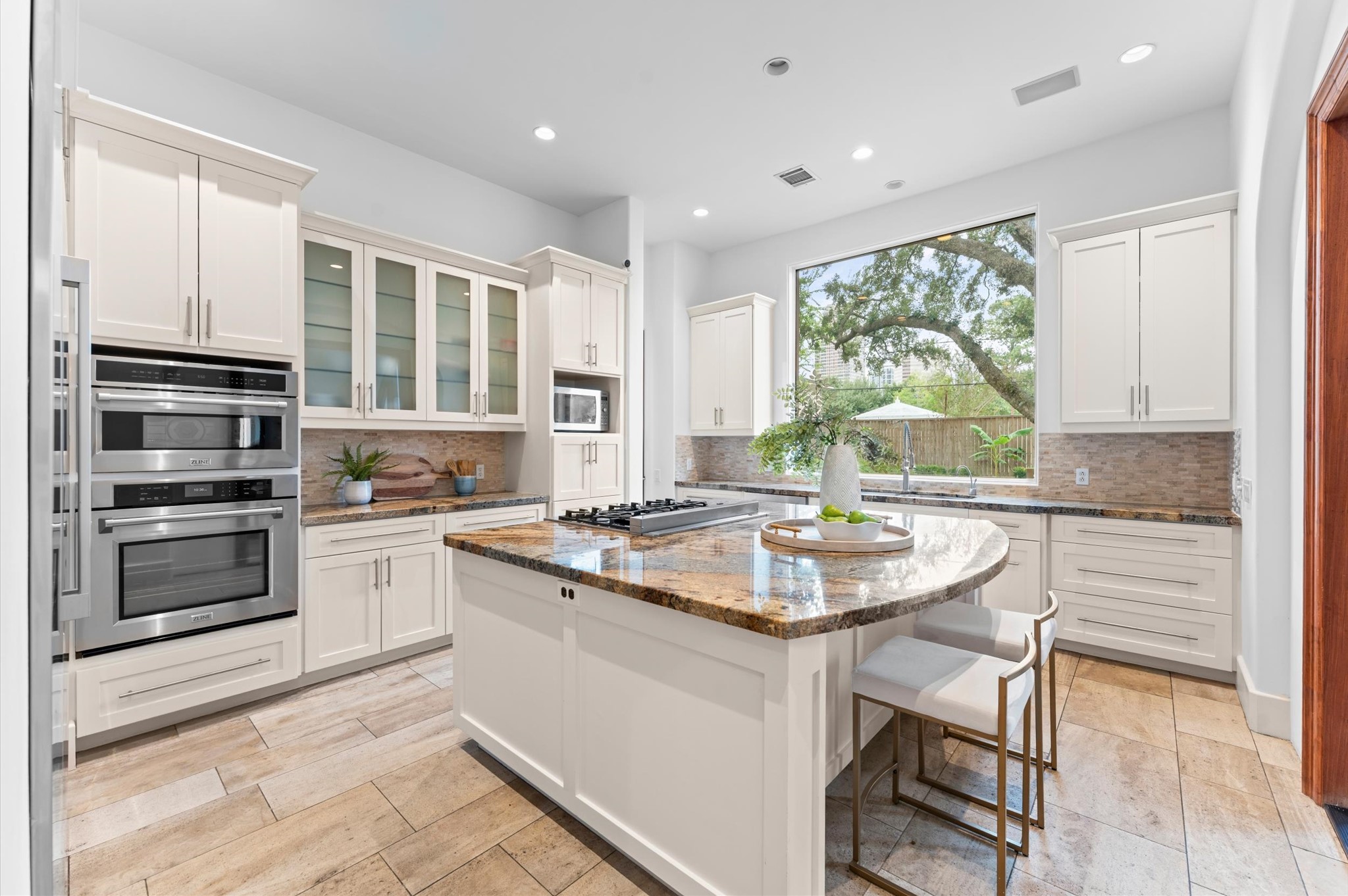 The spacious gourmet kitchen features freshly painted cabinetry, granite countertops, a designer backsplash, travertine flooring, and an expansive island with a gas cooktop. A premium stainless steel appliance suite, including a full-size refrigerator and freezer, complements the kitchen's functional design, while a walk-in pantry provides additional storage.