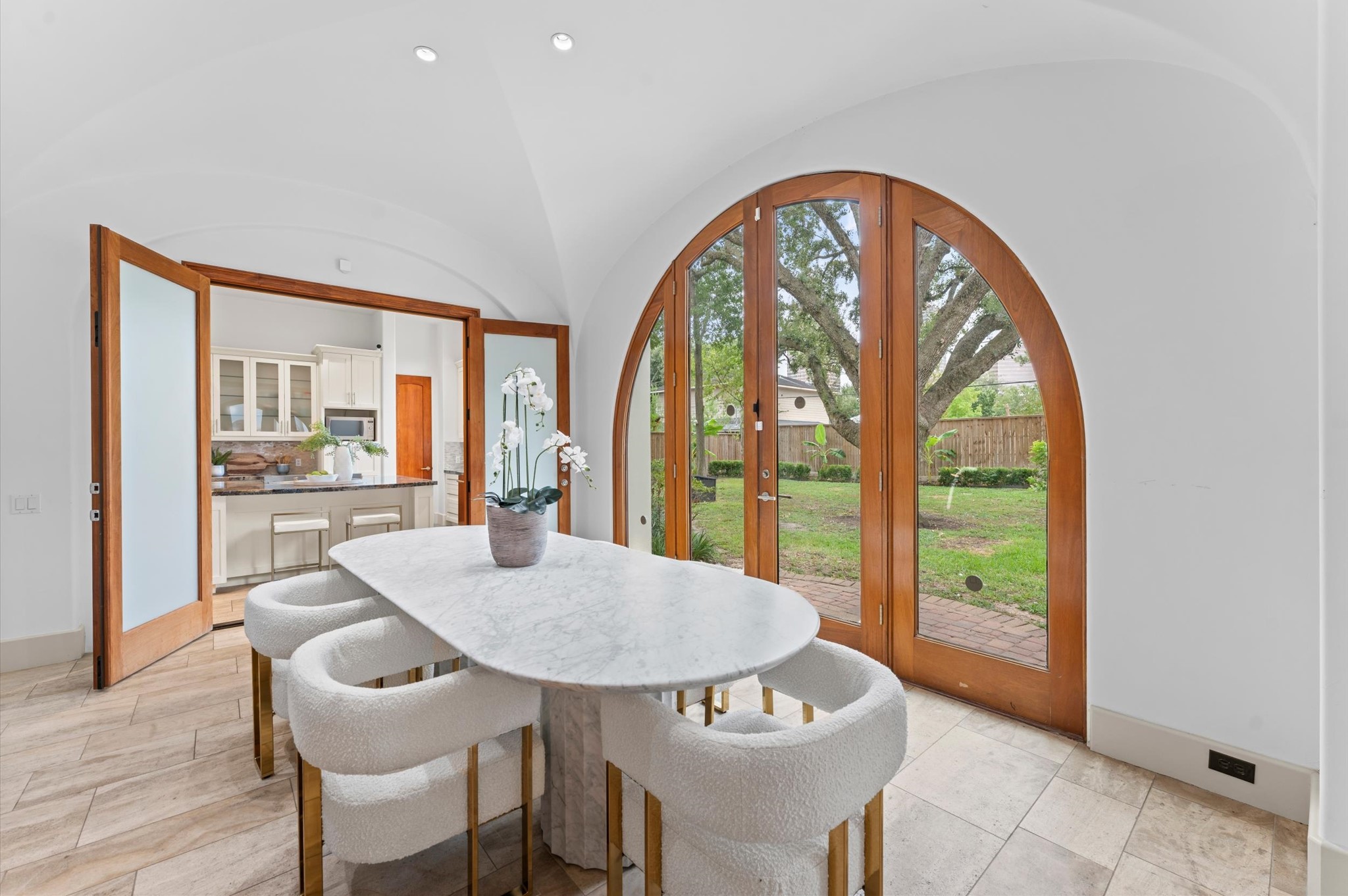 Warm architectural details define the breakfast room, including travertine flooring, plaster walls, a vaulted plaster ceiling, and arched glass doors overlooking the loggia, summer kitchen, pergola, and pool.