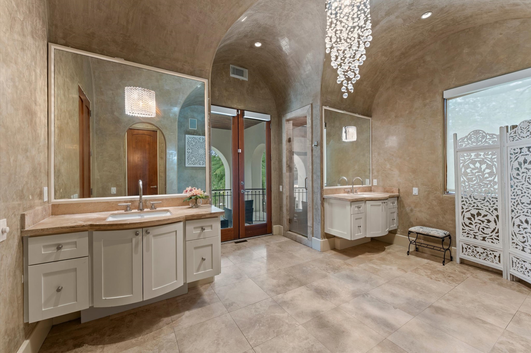 A spa-inspired primary bath features separate floating bombe-style vanities, custom plaster wall finishes, a vaulted ceiling, a glass-enclosed shower, and balcony access.
