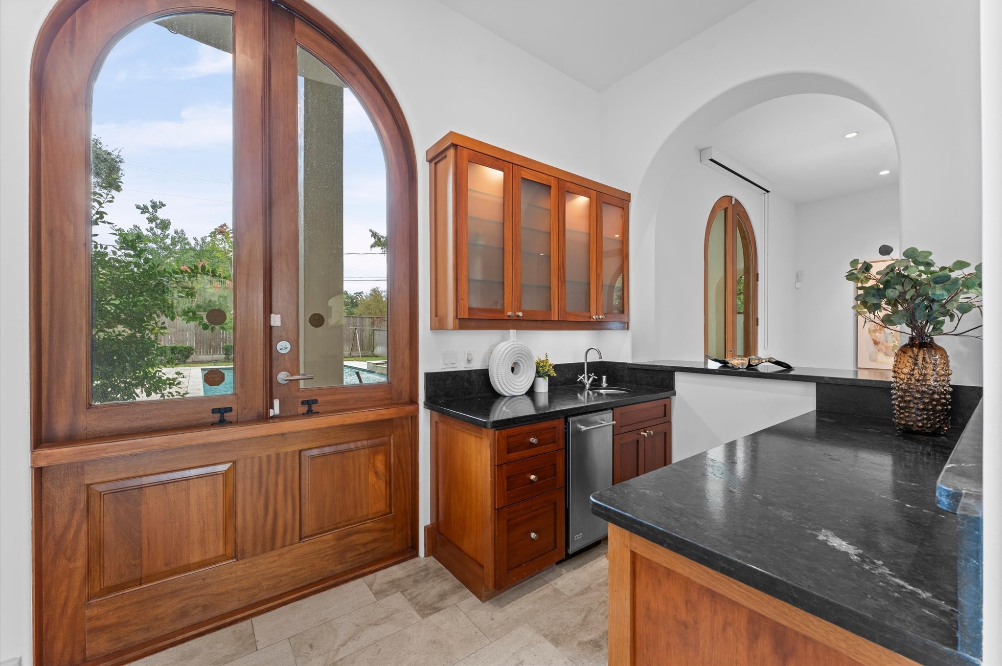 A beautifully appointed wet bar features black marble countertops, a beverage chiller, ice maker, bar sink, and abundant storage. Operable service windows connect the bar to the outdoor kitchen and dining areas.
