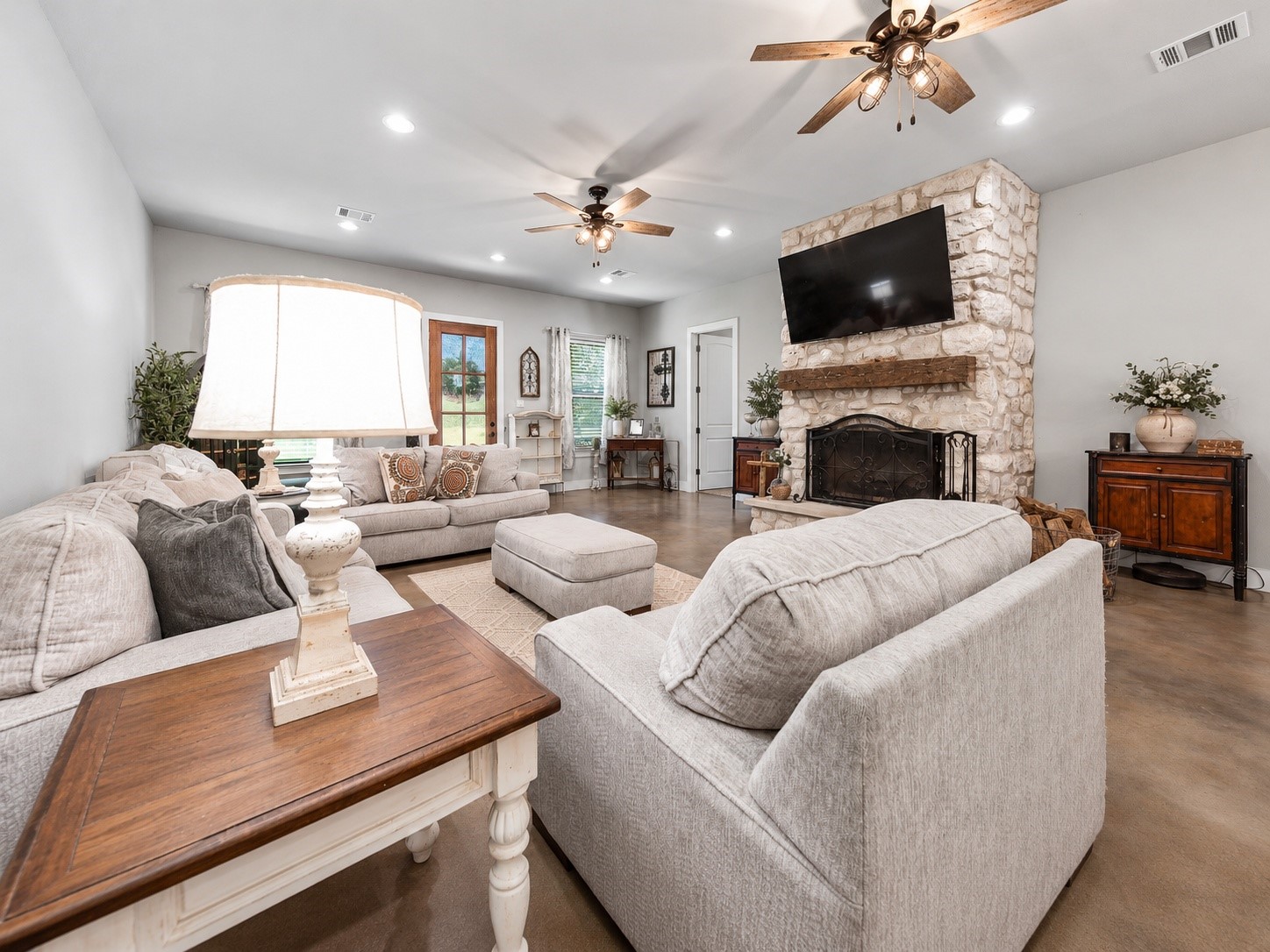 Family room seating area facing the stone fireplace, with ceiling fans and stained concrete floors. This photo may be edited using AI tools.