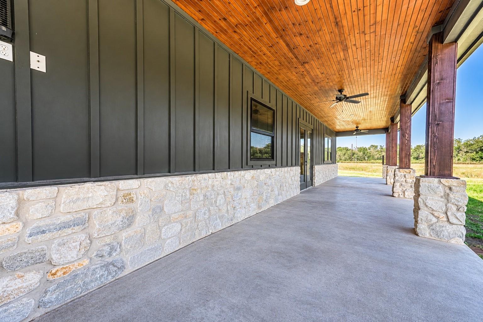 Covered back patio with stained wood ceiling, ceiling fan, stone wainscot and stained concrete floor. This photo may be edited using AI tools.