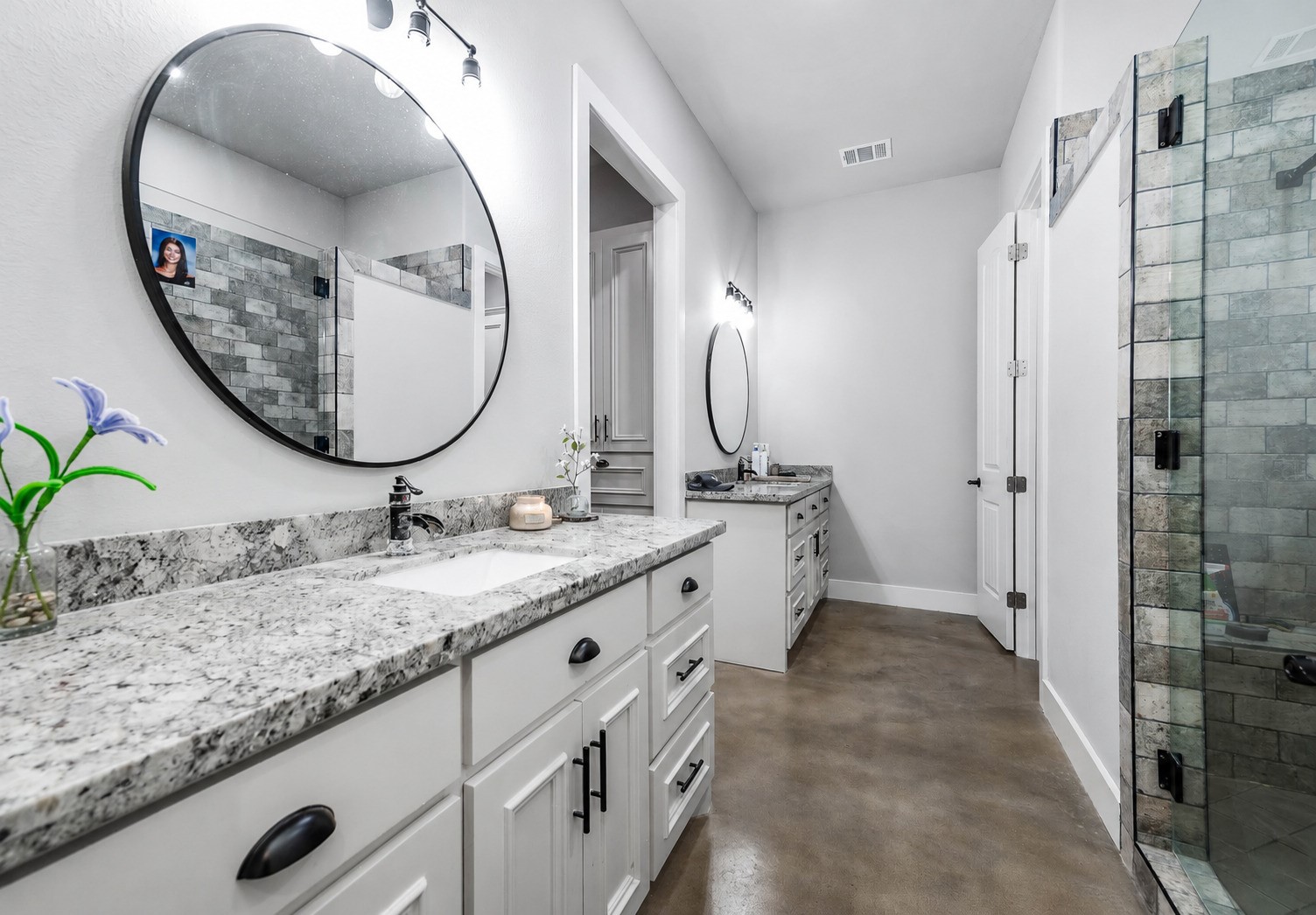 Primary bathroom with double vanity, granite countertops, round mirrors and a glass-enclosed walk-in shower. This photo may be edited using AI tools.