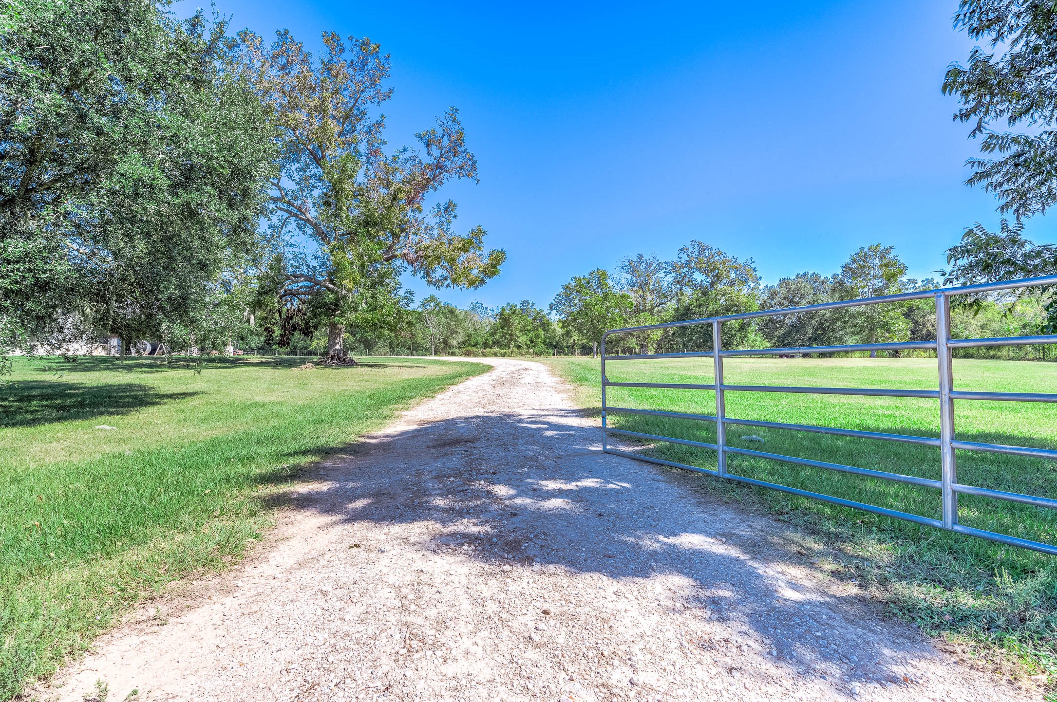Gated gravel drive off FM 524 leading to the home through open pasture and mature trees. This photo may be edited using AI tools.