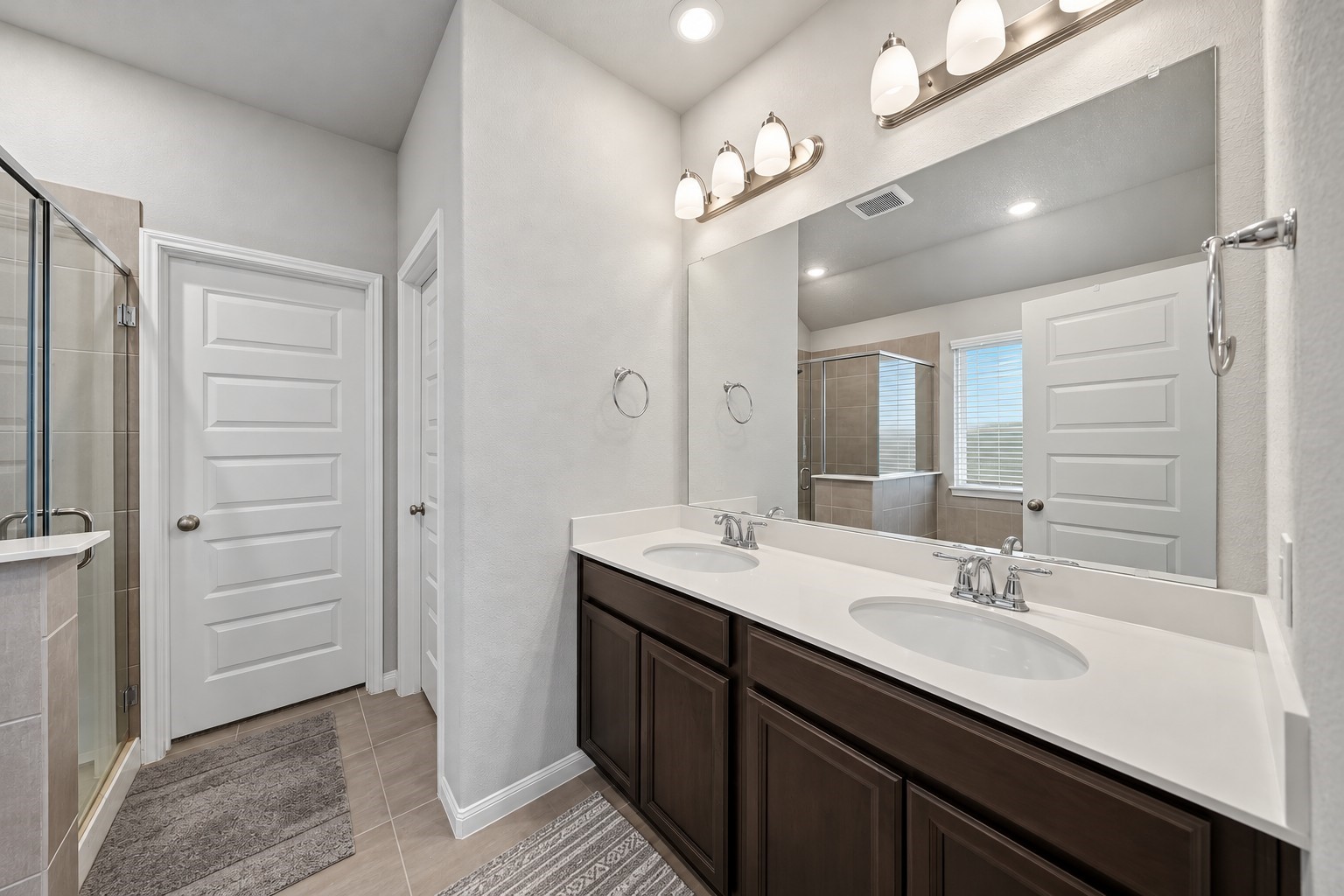 Virtually Enhanced: This spacious primary bathroom features a double-sink vanity with a sleek white countertop, undermount basins, and rich dark cabinetry offering generous storage. A large wall-to-wall mirror, and recessed ceiling lighting create a bright, welcoming atmosphere. Neutral tile flooring complements the soft gray walls and crisp white trim.