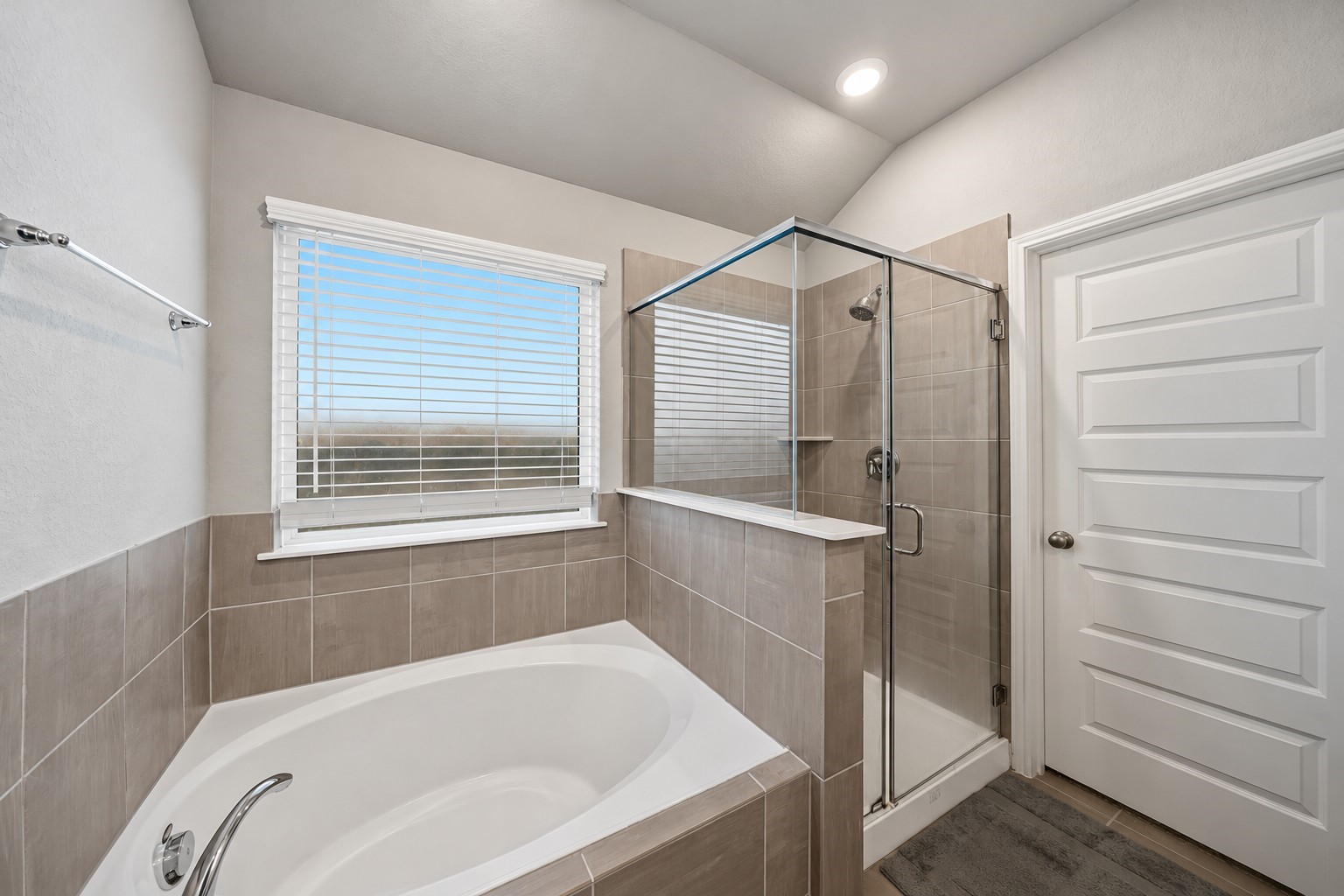 Virtually Enhanced: This spacious primary bathroom features a relaxing soaking tub beneath a large window with white trim and horizontal blinds, allowing natural light to brighten the space. A separate glass-enclosed walk-in shower with a chrome-framed door provides added convenience and functionality. Neutral-toned tile surrounds complement the walls, while the vaulted ceiling enhances the open, airy atmosphere.