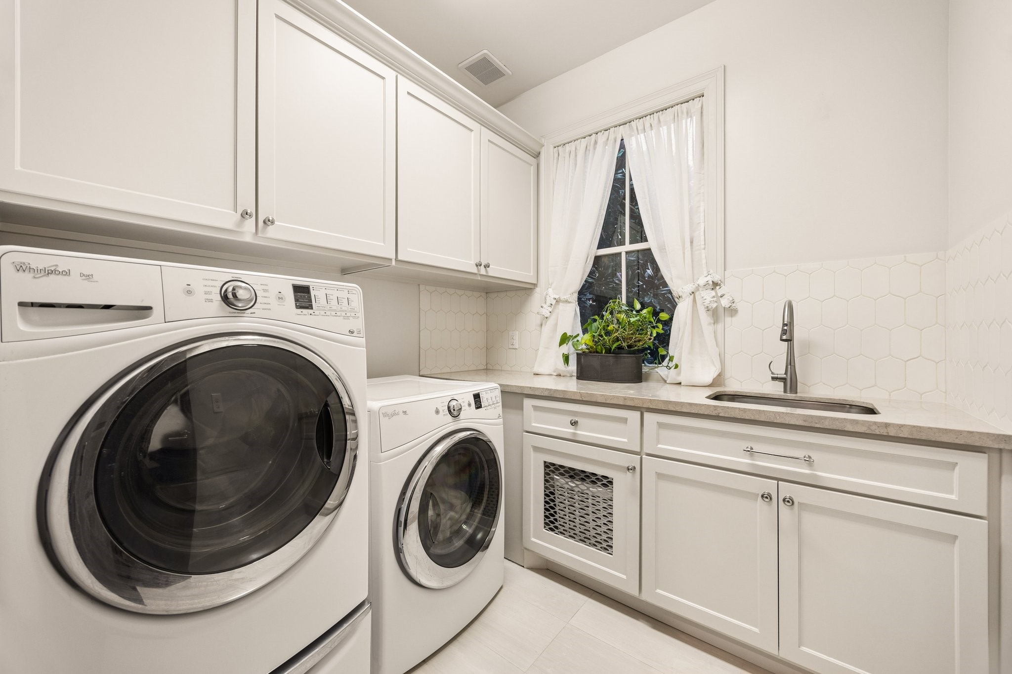 Bright, beautifully appointed laundry room featuring Whirlpool washer and dryer, custom cabinetry, generous counter space, utility sink, and elegant tile backsplash.