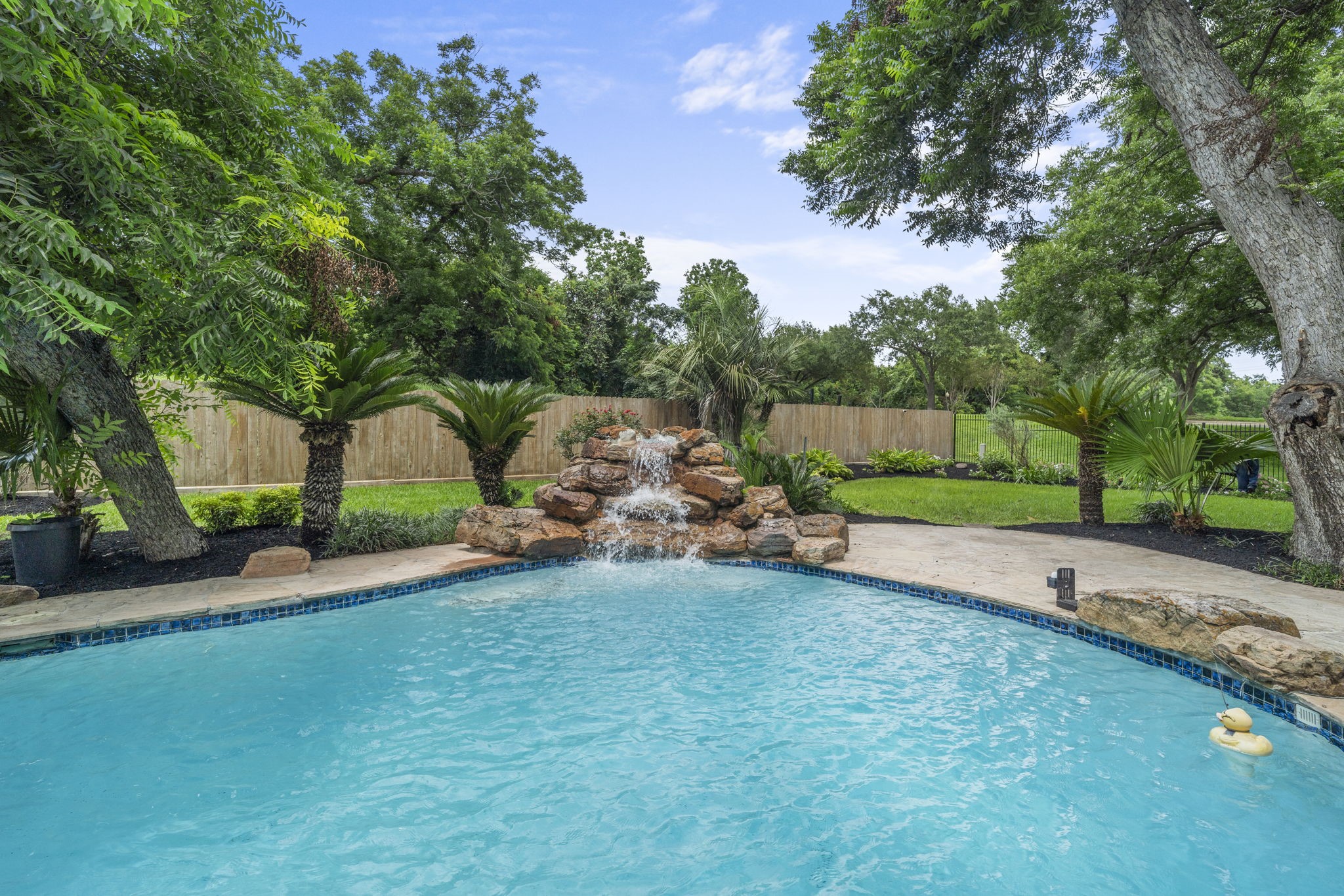 A cascading rock waterfall creates a beautiful focal point, adding movement & sound to the pool setting while mature trees frame the backyard.