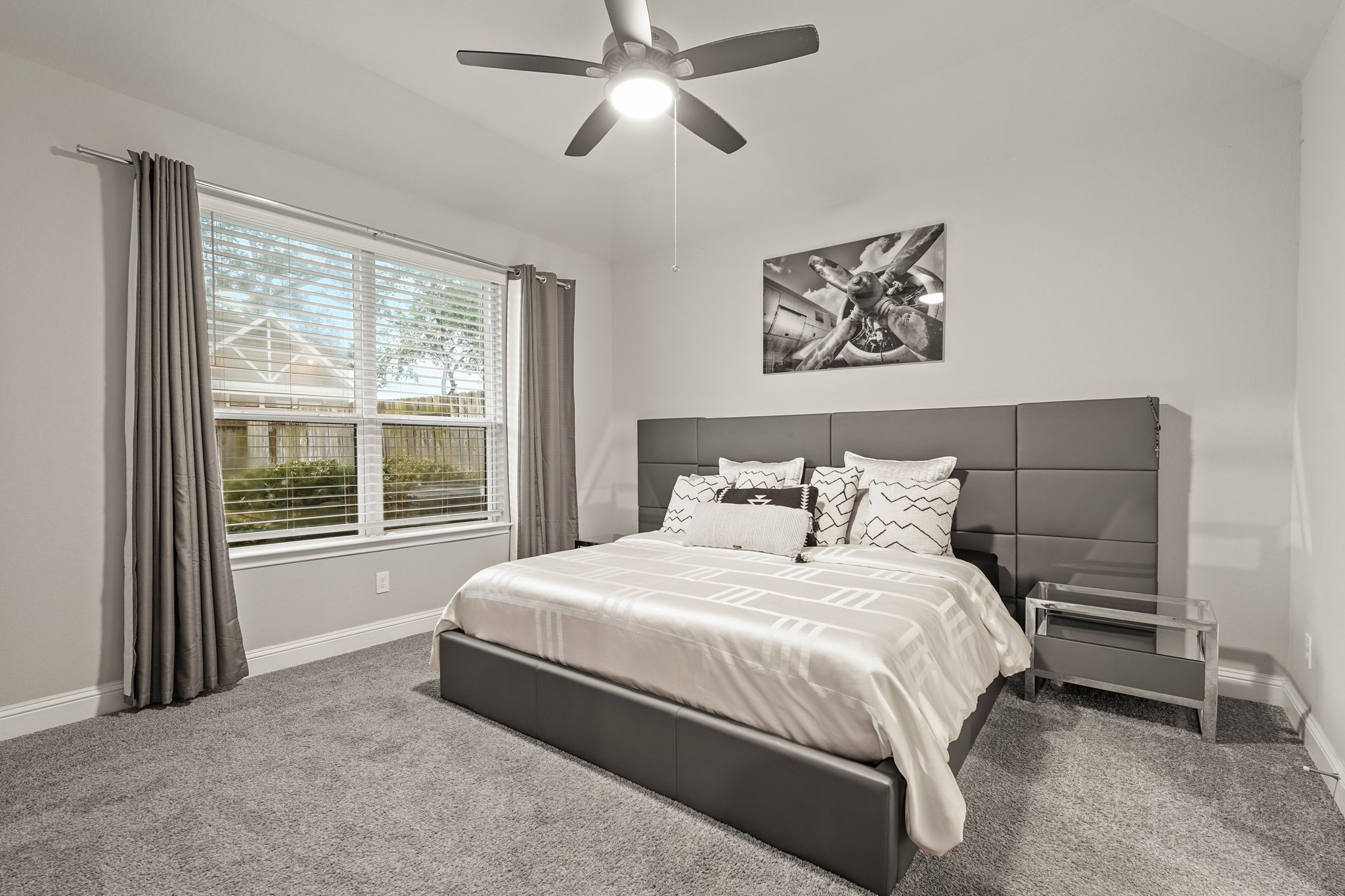 A bright and welcoming primary bedroom, showing off a great layout for a main suite. The room has a modern, cohesive look with its neutral grey walls, white trim, and plush dark carpeting. A large double window provides a nice view of the outside and lets in great natural light. The high vaulted ceiling adds an open, airy feel, making this primary bedroom a comfortable and inviting retreat at the end of the day.