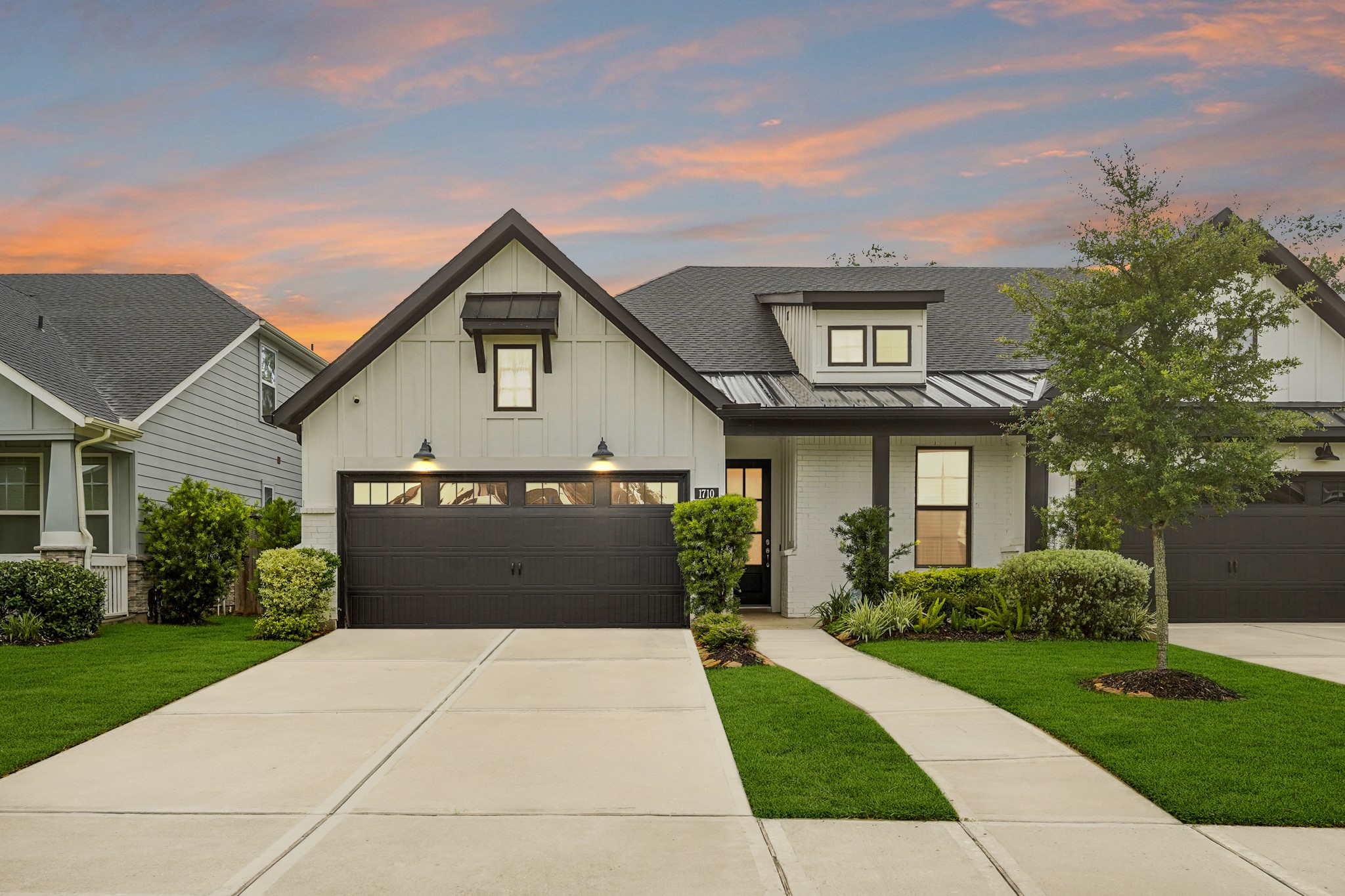 This front view of the home, highlights its modern curb appeal. The exterior features crisp white board-and-batten siding paired with white painted brick, all beautifully contrasted by a dark shingle roof and deep black trim. A wide concrete driveway leads up to a spacious two-car garage with a matching black garage door.