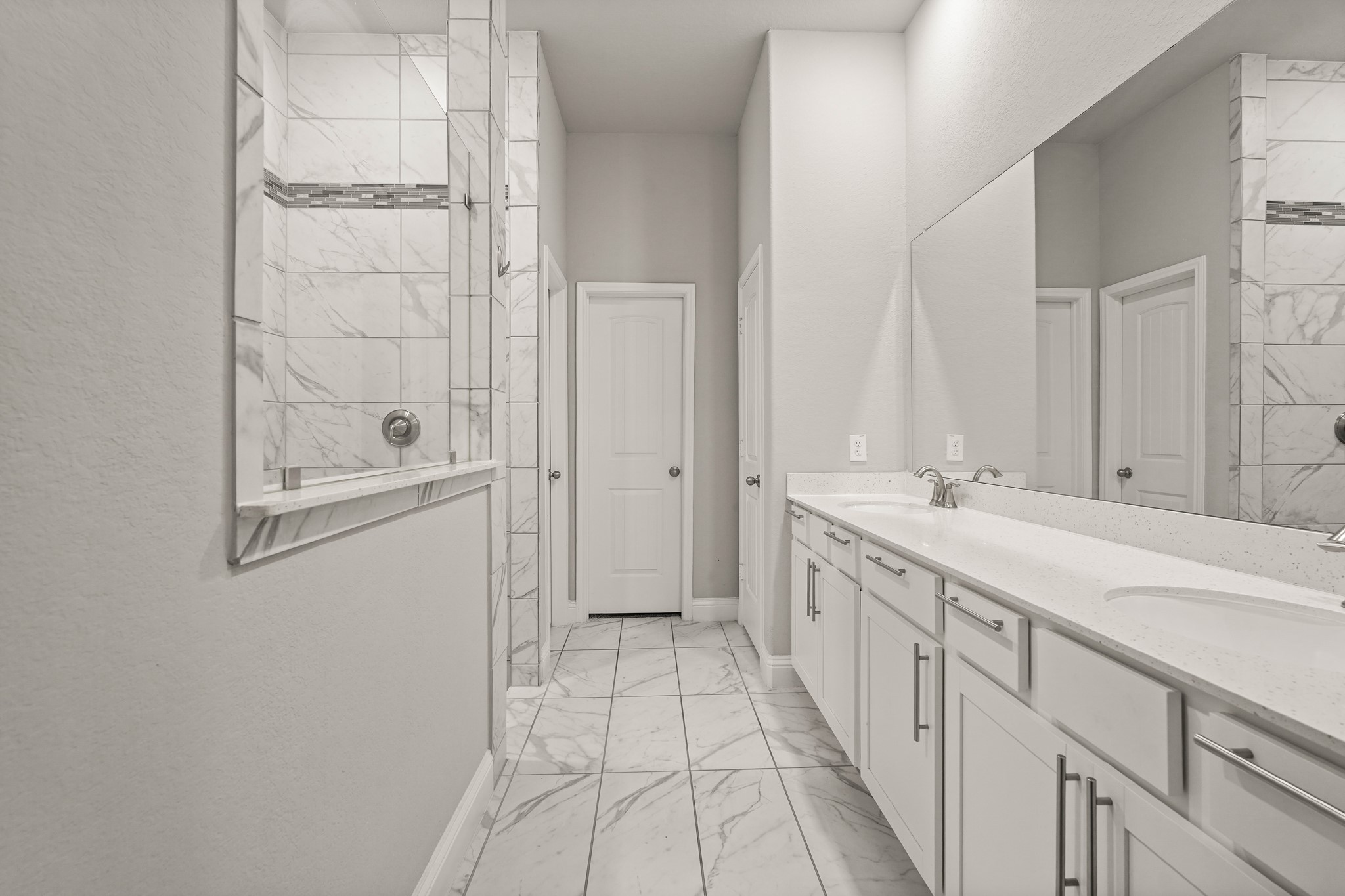 This spacious main bathroom features a long, functional layout with clean finishes. On the right, a wide double vanity offers plenty of storage with white cabinets, long silver handles, and a bright white countertop with dual sinks under a massive wall mirror. On the left, the walk-in shower is styled with large-format marble-look wall tile and a dark mosaic accent strip.