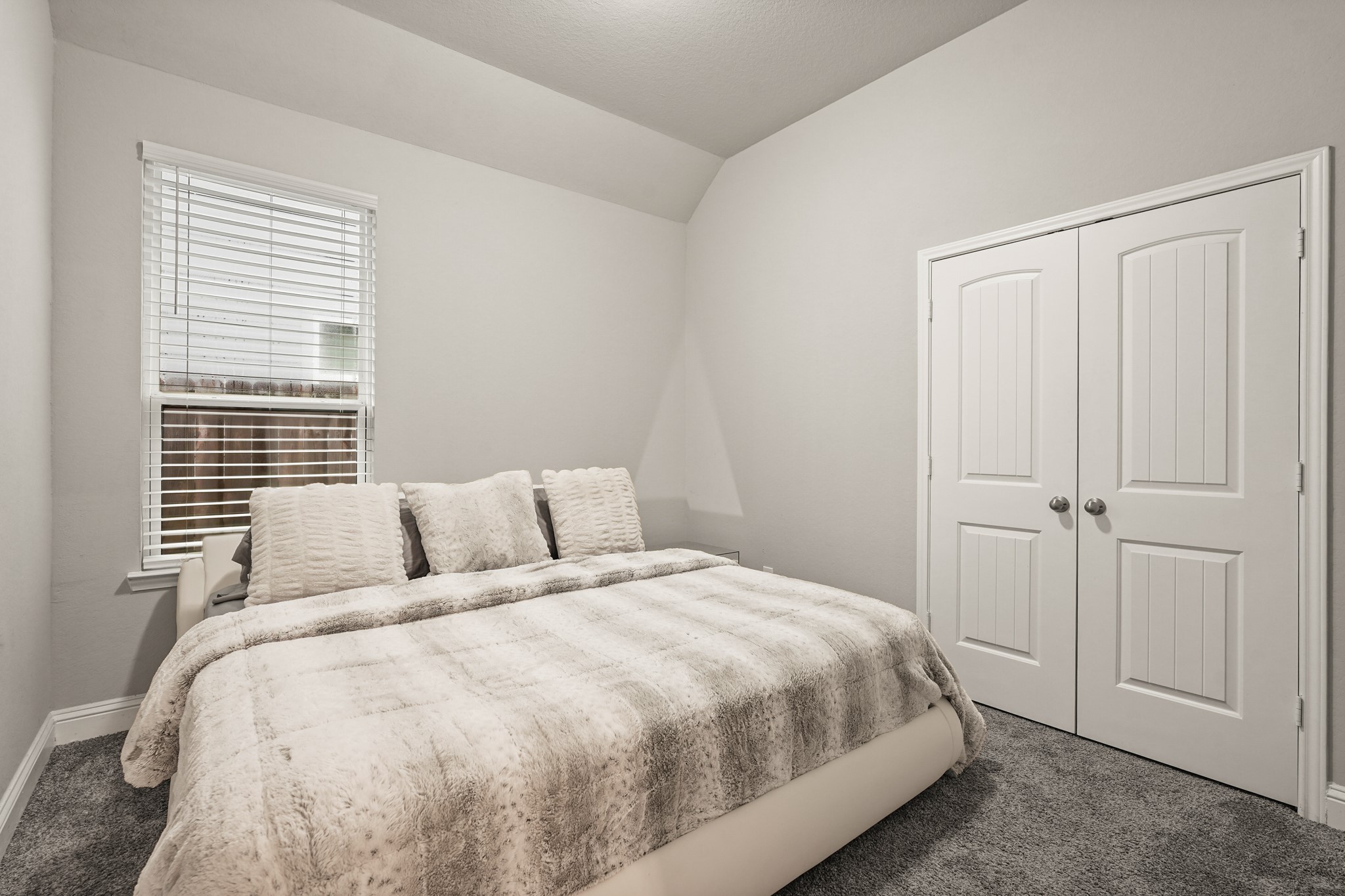 This inviting bedroom features deep grey carpeting for a warm, comfortable atmosphere. A large window with built-in blinds provides excellent natural light, while the vaulted ceiling detail adds architectural interest and vertical space. Double closet doors with classic raised panel details complete the room, offering ample storage space.