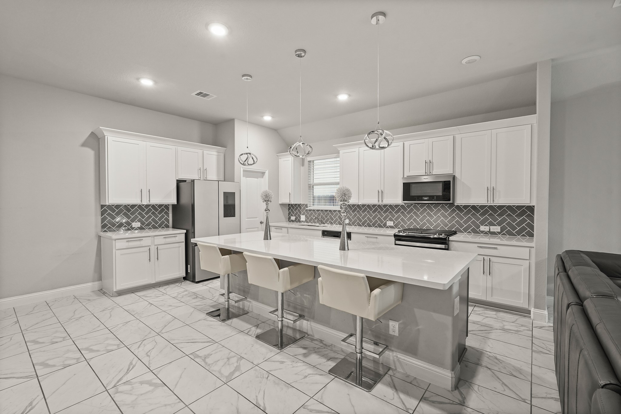 This bright, contemporary kitchen features an expansive island with seating and a seamless white polished countertop. The design includes timeless white cabinetry with sleek silver hardware, a striking grey herringbone tile backsplash, and matching solid surfaces. Modern spherical pendant lights, recessed ceiling lights, and elegant marble-look tile flooring beautifully complete the space. This bright, contemporary kitchen features an expansive island with seating and a seamless white polished countertop. The design includes timeless white cabinetry with sleek silver hardware, a striking grey herringbone tile backsplash, and matching solid surfaces. Modern spherical pendant lights, recessed ceiling lights, and elegant marble-look tile flooring beautifully complete the space.