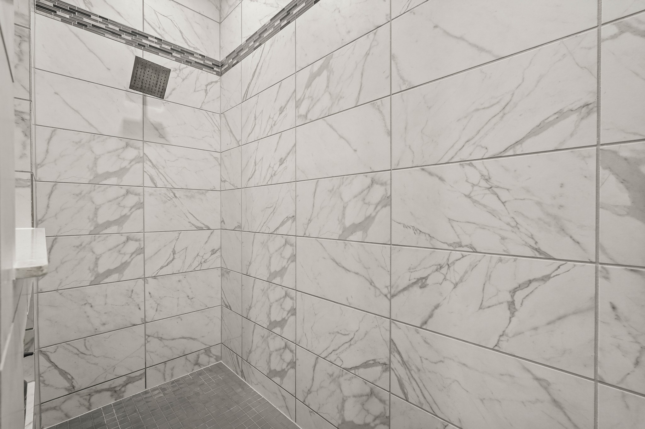 This detailed look inside the custom walk-in shower, highlights its clean and modern design. The spacious enclosure is fully lined with large-format wall tiles featuring a bright white and grey marble pattern. A stylish horizontal mosaic tile accent strip runs along the upper perimeter of the walls.