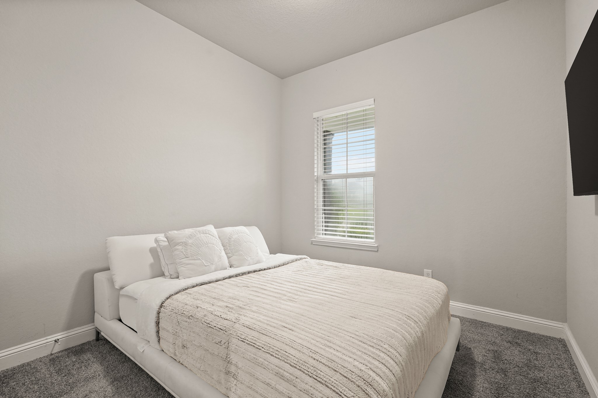 This cozy secondary bedroom offers a bright and inviting atmosphere with soft neutral tones and abundant natural light. The room features plush carpeting, crisp white trim, creating a clean and modern aesthetic. A large window with blinds allows natural light to fill the space while offering views of the outdoors.