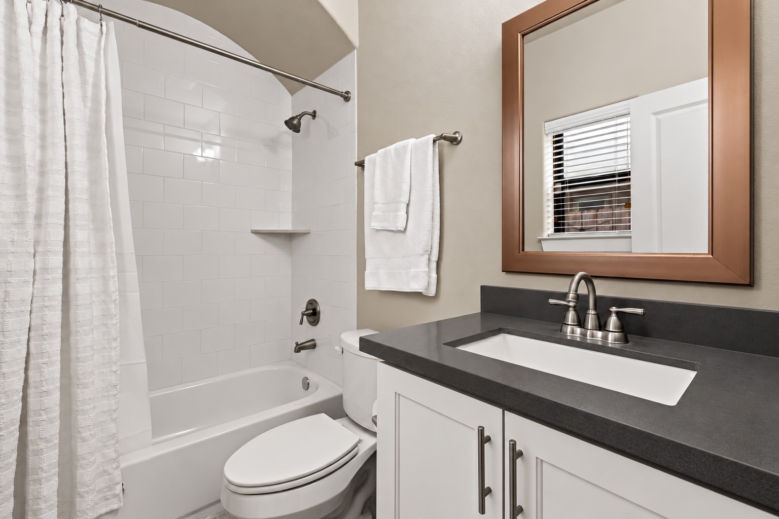A dark vanity counter strikes a refined contrast against white cabinetry and pale walls. The adjoining tub-and-shower combination completes a comfortable full bath. A dark vanity counter strikes a refined contrast against white cabinetry and pale walls. The adjoining tub-and-shower combination completes a comfortable full bath.