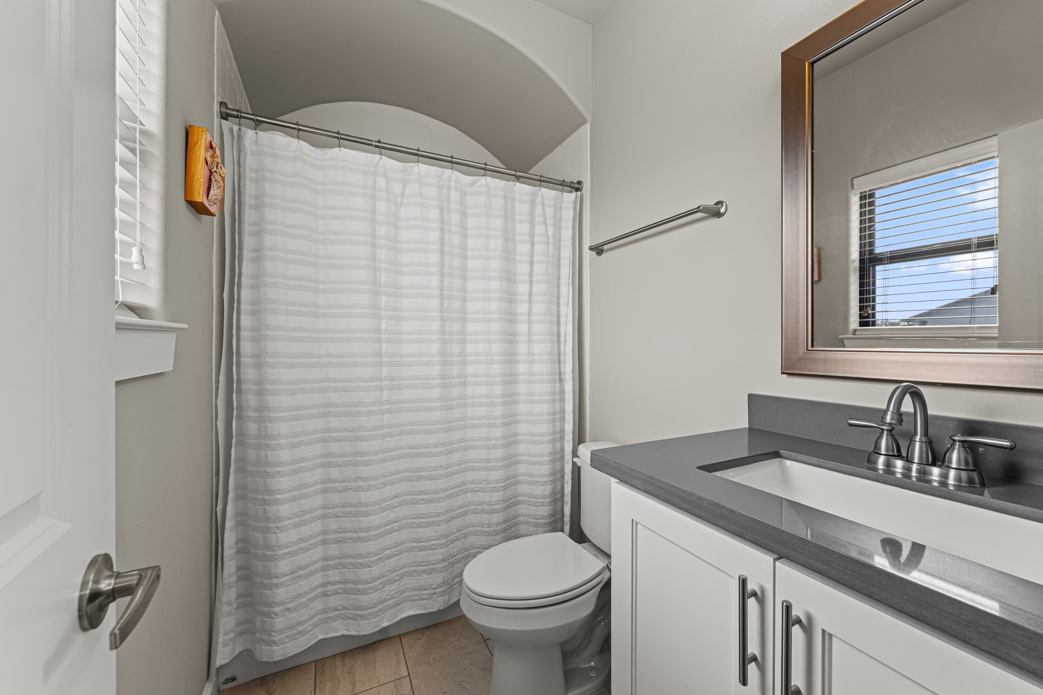 An arched opening frames the tub-and-shower area with a graceful architectural flourish. A dark vanity counter and nearby window balance rich contrast with natural light.