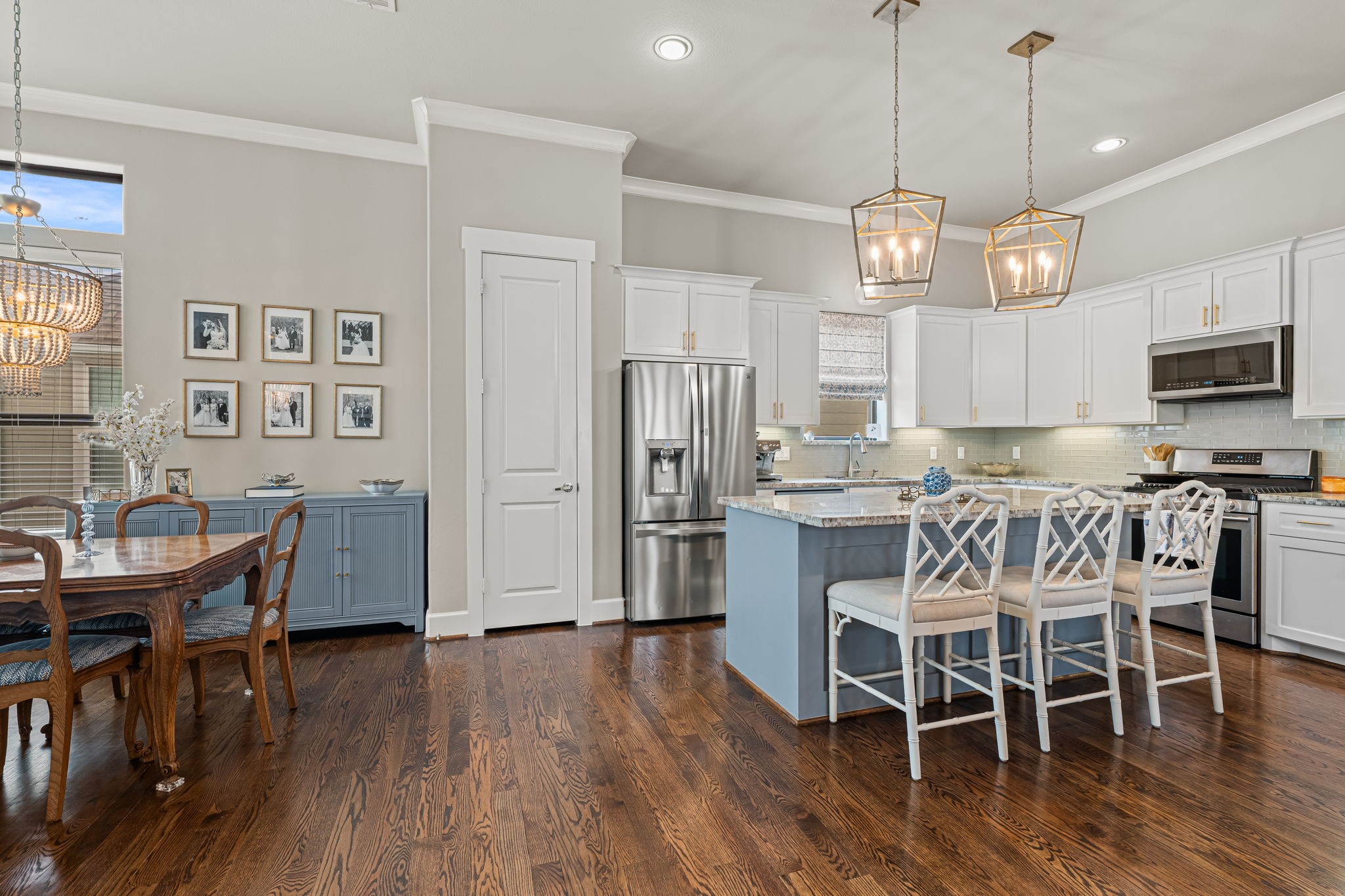 A blue island brings a distinctive note of color to the kitchen beneath a pair of designer lantern pendants. White cabinetry, stainless-steel appliances, and a softly textured backsplash complete the inviting palette. Note the custom window treatment.