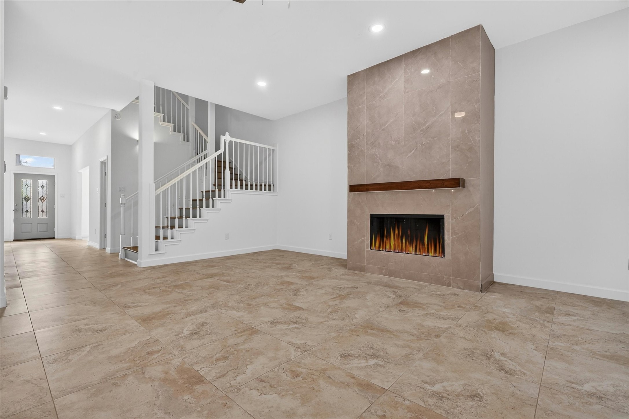 Spacious open-concept living room featuring soaring ceilings, oversized tile flooring, recessed lighting, and a stunning floor-to-ceiling tiled fireplace with modern wood mantle. Elegant staircase with wood accents creates a grand focal point, while the bright neutral finishes make this home move-in ready and perfect for entertaining.