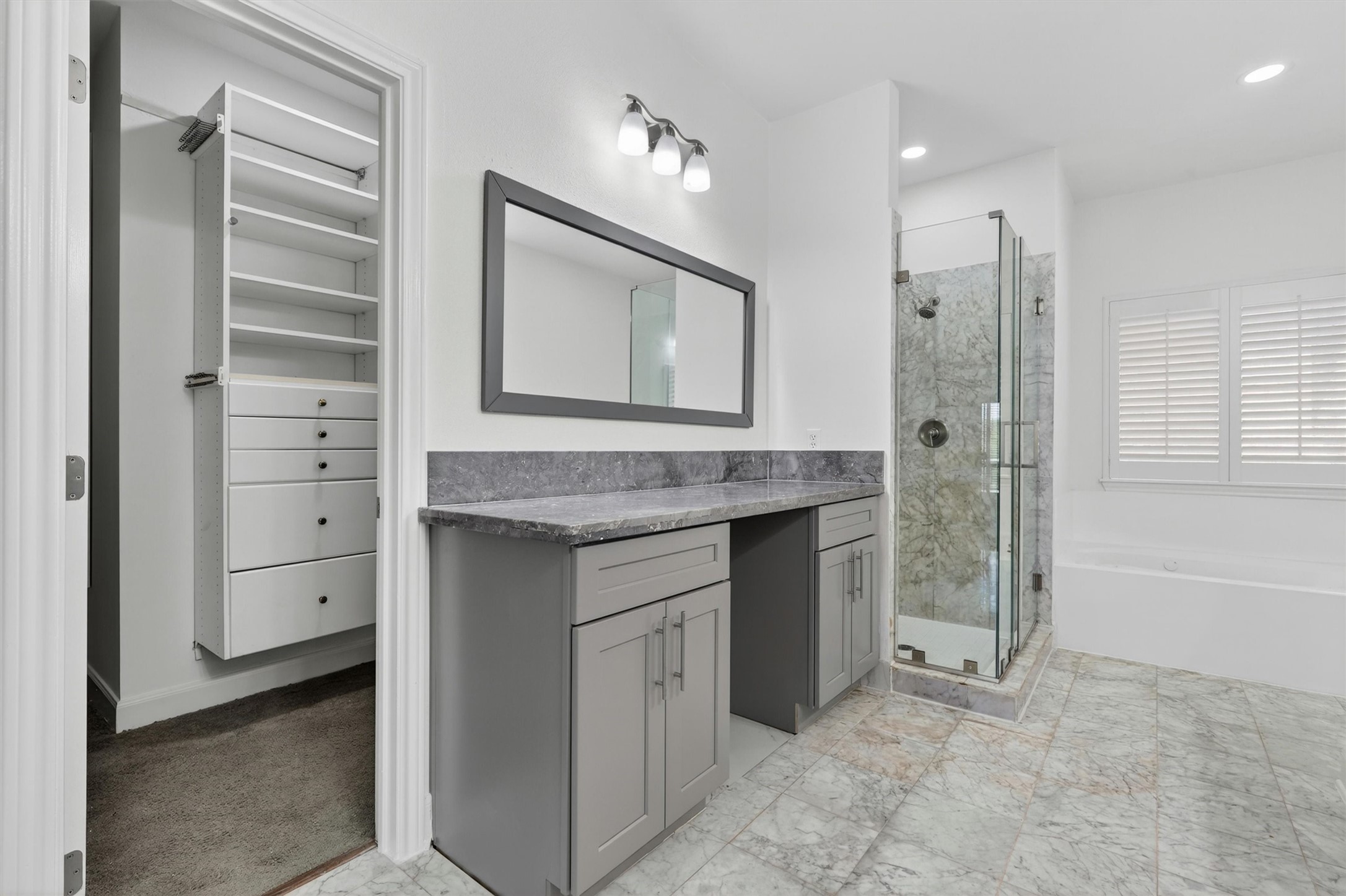 Luxurious primary bathroom featuring marble-look tile flooring, oversized vanity with granite countertops, framed mirror, and designer lighting. Spa-inspired walk-in shower with frameless glass enclosure, soaking tub, plantation shutters, and custom walk-in closet with built-in shelving complete this elegant retreat.
