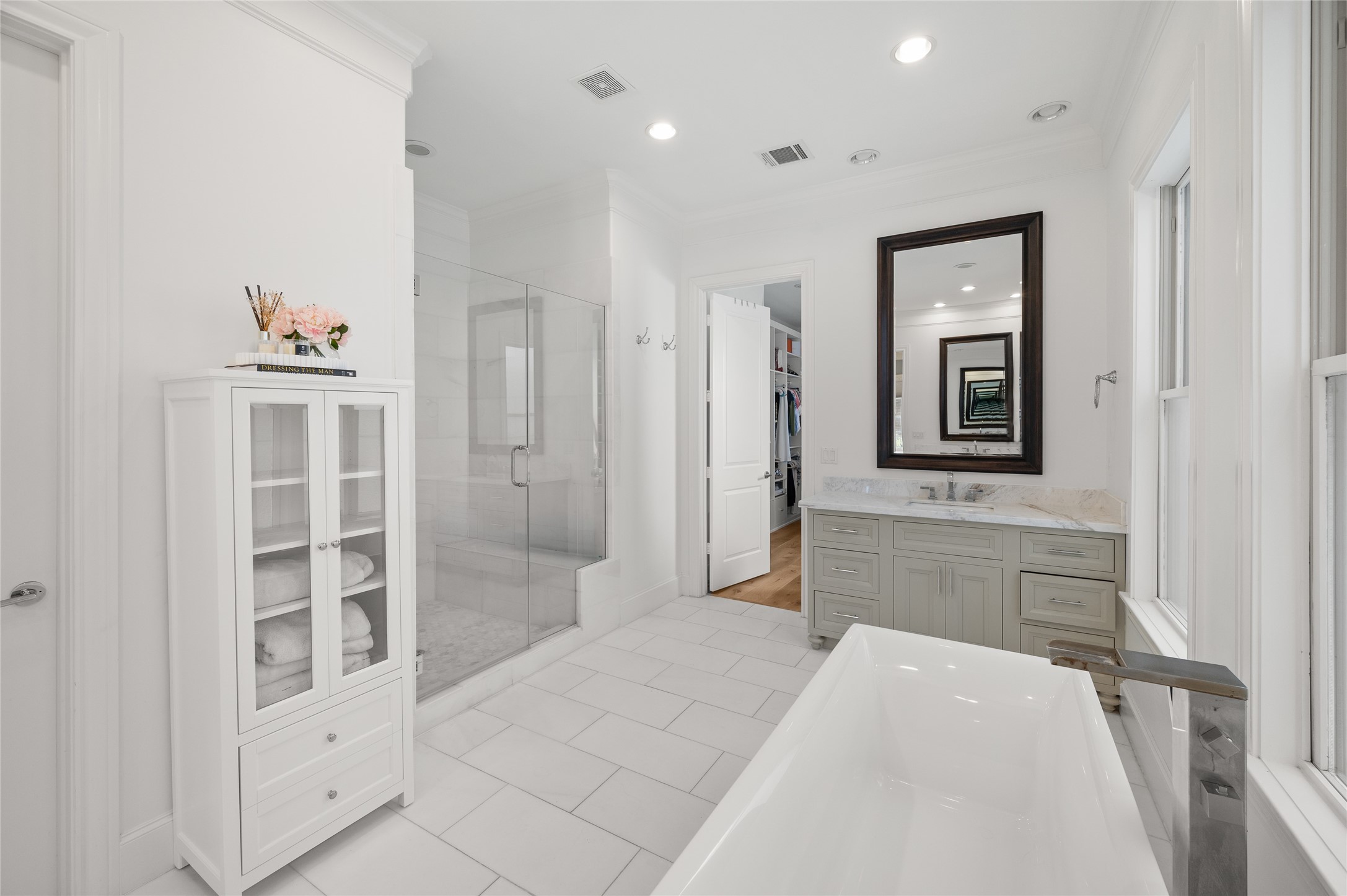 Freestanding soaking tub positioned beneath three windows, with marble-topped vanity and elegant white tile flooring.