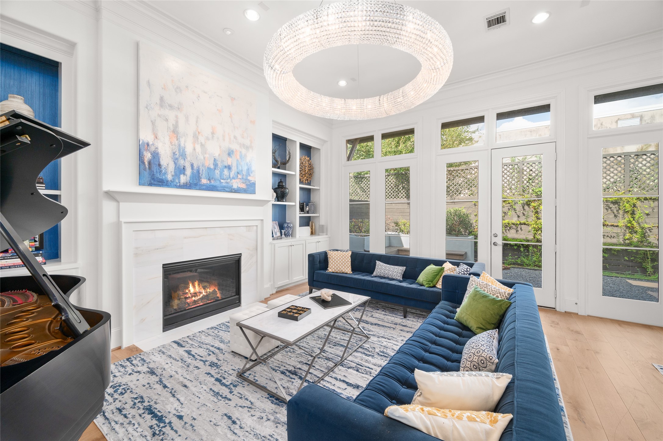 Sunlit living room with a marble-clad fireplace, dramatic circular chandelier, and a wall of windows and French doors opening to the courtyard.