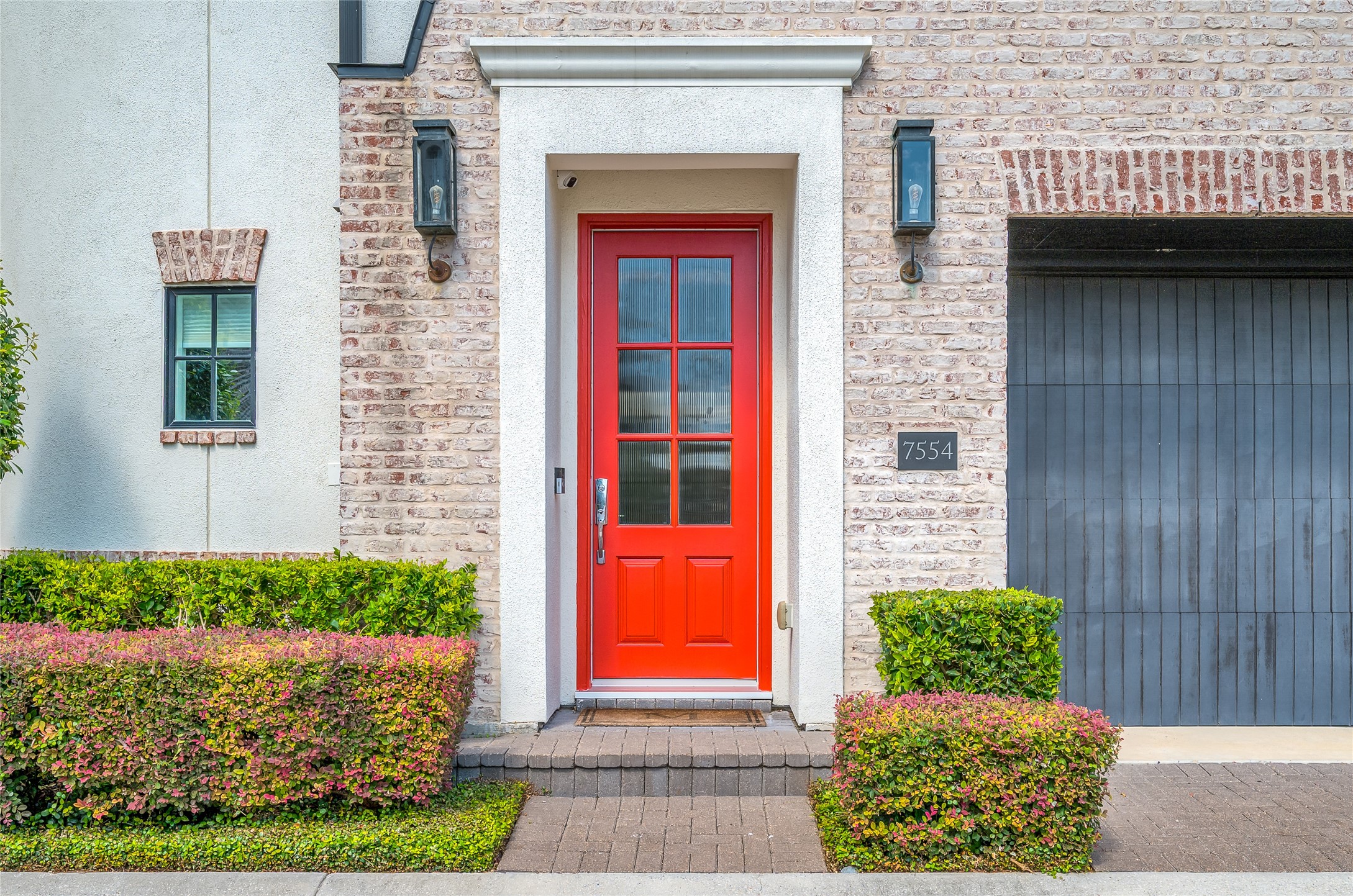 A striking red entry door makes a memorable first impression, framed by gorgeous brickwork, classic lantern-style sconces, and meticulously manicured landscaping.