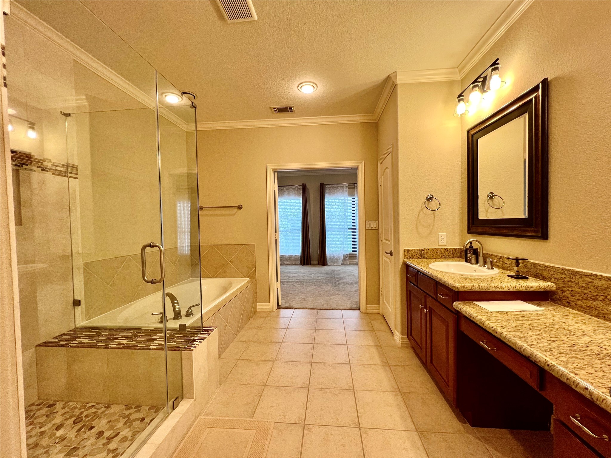This spacious bathroom features a glass-enclosed shower and a separate bathtub, with elegant tile work throughout. It includes a large granite countertop with dual sinks and ample cabinet space.