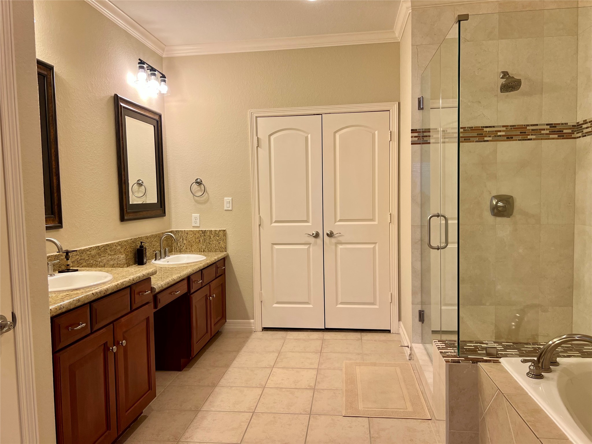 This master bathroom features a spacious layout with dual sinks, granite countertops, and wooden cabinets. It includes a glass-enclosed shower and a separate bathtub, offering a blend of functionality and elegance. There is a closet at the end of the bathroom