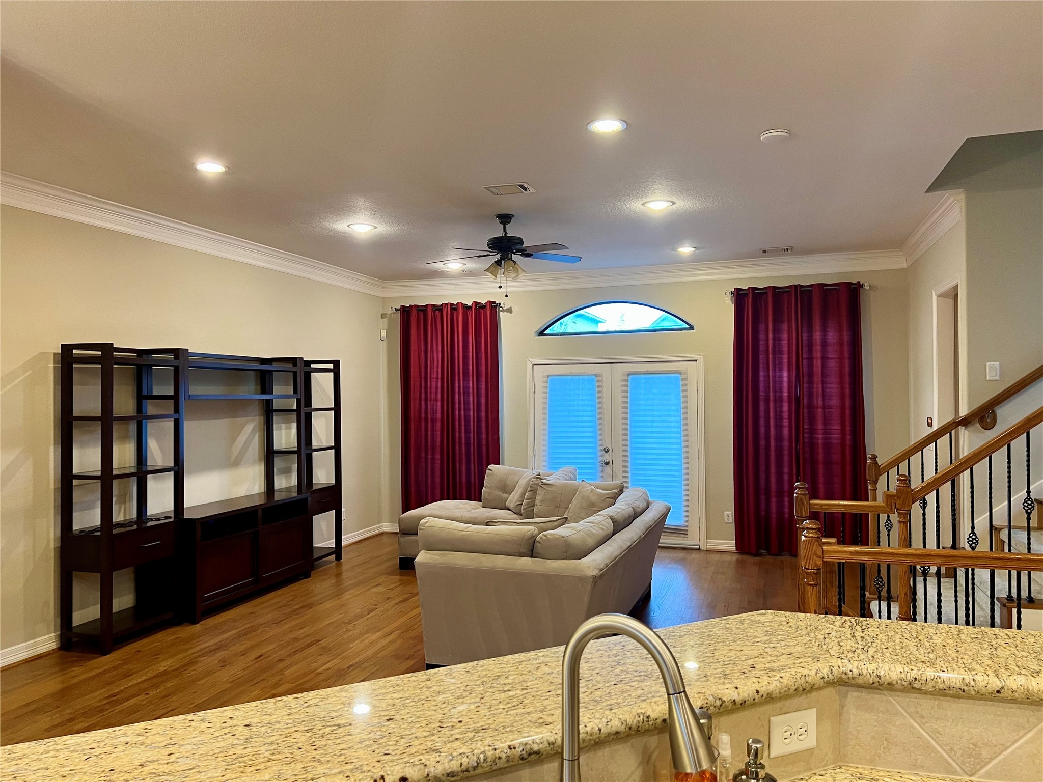 Spacious living room with hardwood floors, featuring a cozy sectional sofa and a large entertainment unit. The room is well-lit with recessed lighting and a ceiling fan. French doors with red curtains lead outside