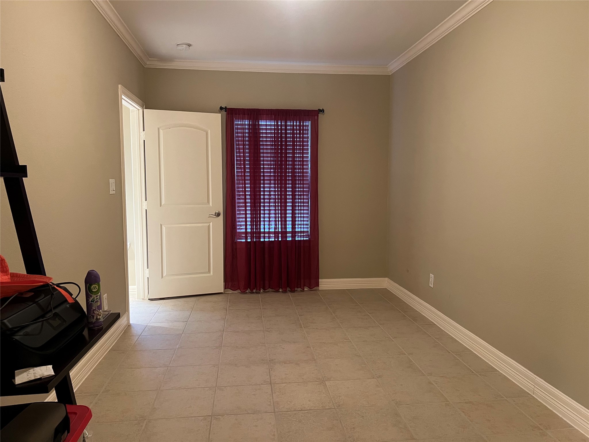 This room features neutral walls, tiled flooring, and a window with red curtains allowing natural light. It has a white door and crown molding, providing a clean and simple space. This room features neutral walls, tiled flooring, and a window with red curtains allowing natural light. It has a white door and crown molding, providing a clean and simple space.