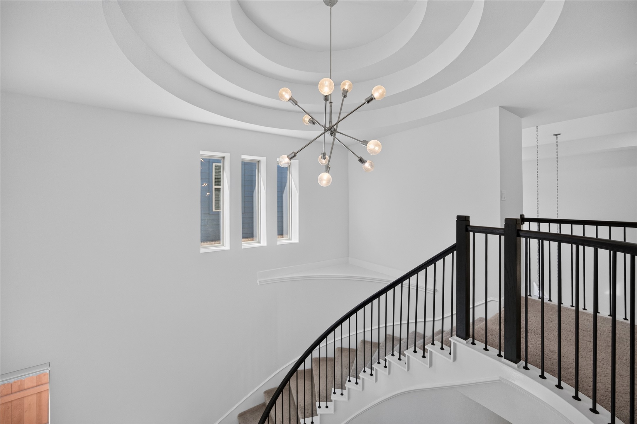 The upstairs landing provides a close view of the homes dramatic tiered ceiling and contemporary chandelier. Curved iron railing follows the staircase and overlooks the foyer below. A trio of windows introduces natural light and emphasizes the architectural detail of this central space.