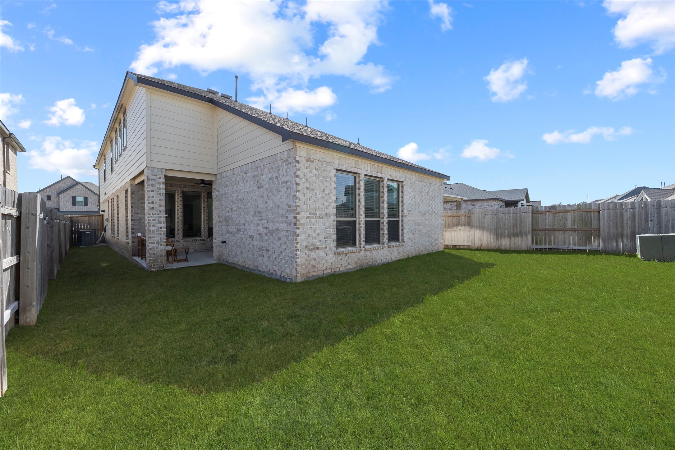 This rear elevation highlights the homes light brick exterior, upper siding and covered patio tucked beneath the main roofline. Large rear windows connect the interior to the backyard, while the surrounding lawn provides usable outdoor space. Wood fencing encloses the yard along the neighboring properties.