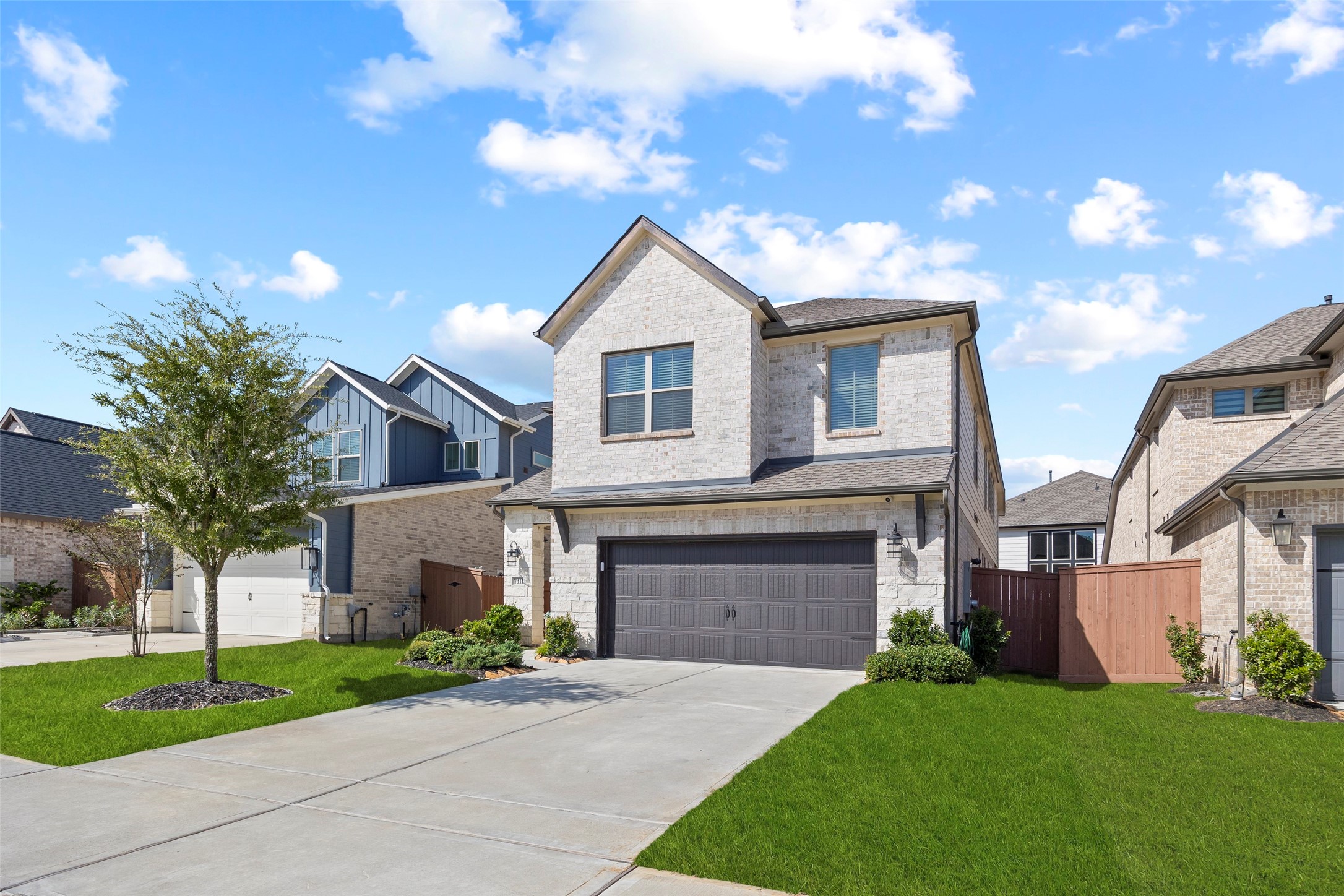 This angled view highlights the homes architectural lines, light brick exterior and contrasting garage door. A double-width driveway provides access to the attached two-car garage, while the front lawn, planting beds and established tree soften the approach to the covered entry.