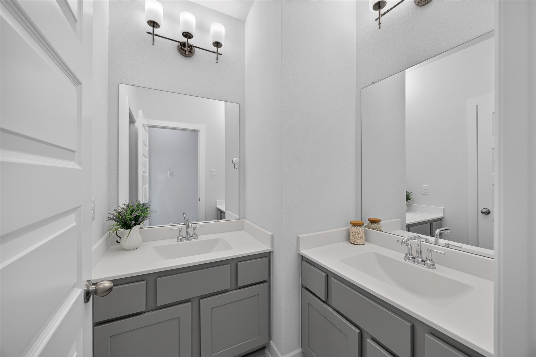 Two separate vanity areas provide individual sinks, mirrors and cabinet storage in this secondary bathroom. The arrangement creates distinct spaces for daily routines, with light countertops and gray cabinetry maintaining a cohesive appearance. An adjoining doorway connects the vanity space to the bathing area.