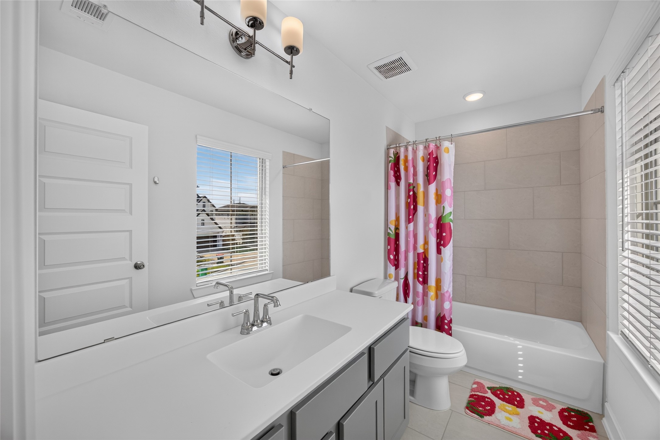 This secondary bathroom includes a long vanity with cabinet storage, a large mirror and a window for natural light. The tub-and-shower combination features a tiled surround, offering flexibility for daily bathing. Light walls and gray cabinetry coordinate with the finishes used elsewhere in the home.