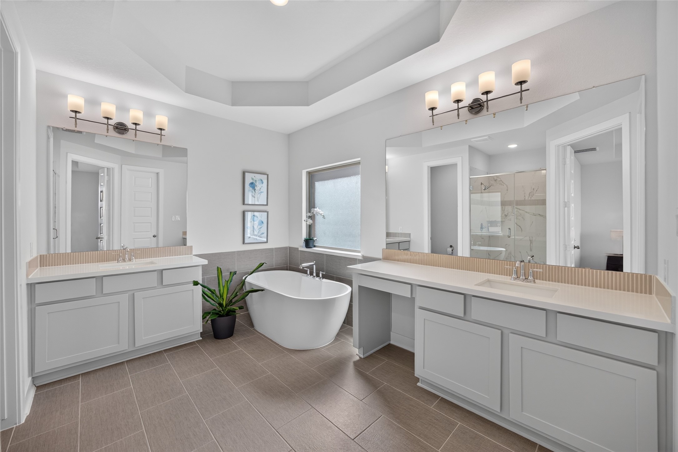The primary bathroom features two separate vanity areas with gray cabinetry, broad mirrors and decorative lighting. A freestanding soaking tub sits beneath the window between the vanities, creating a focal point. A tiered ceiling adds architectural detail, while cabinetry and counter space support everyday routines.