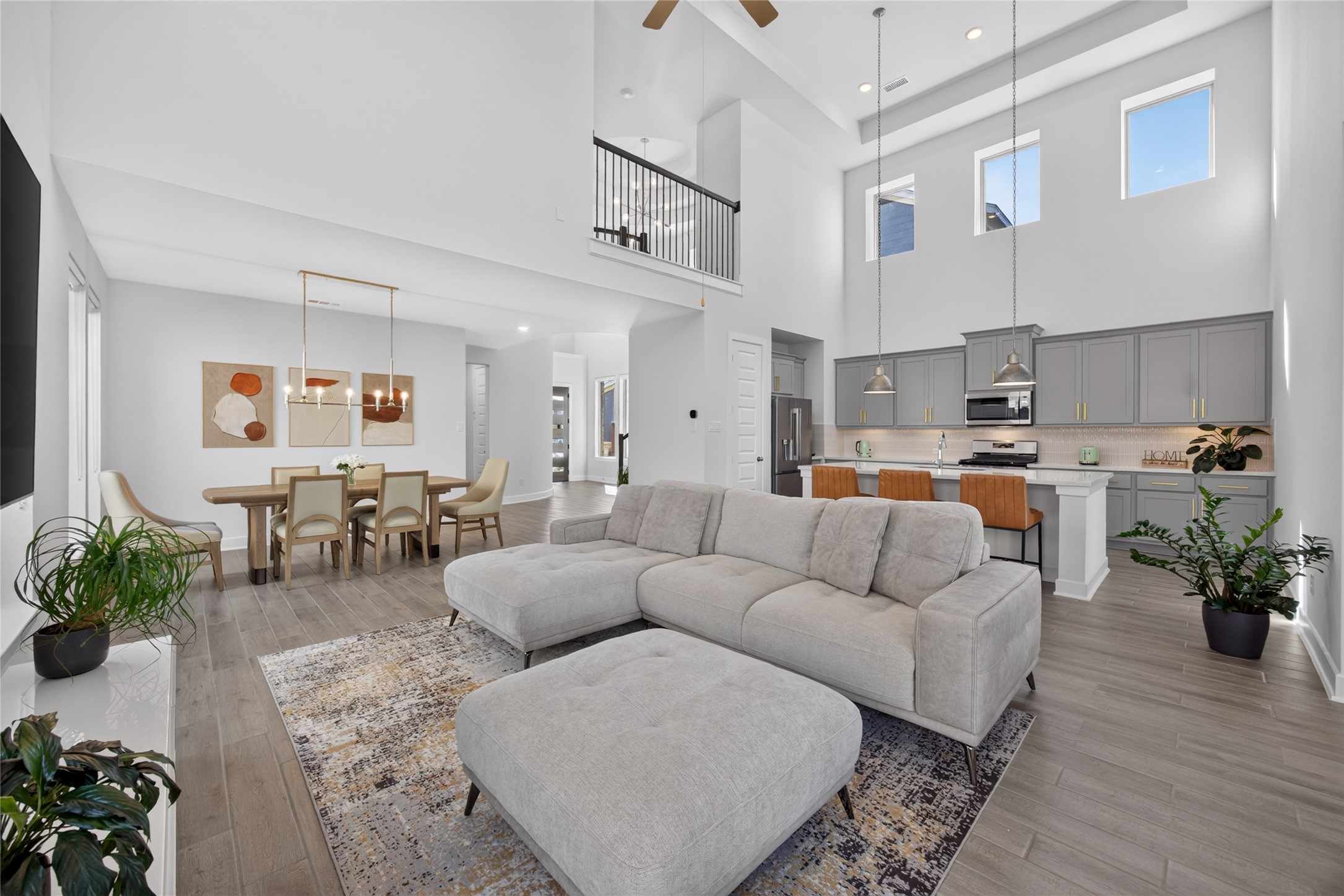 The family room, dining area and island kitchen share an open layout designed for gathering. Two levels of windows accentuate the high ceilings, while the upstairs railing adds architectural interest above. Continuous wood-look flooring connects the spaces and allows furniture to define each area.