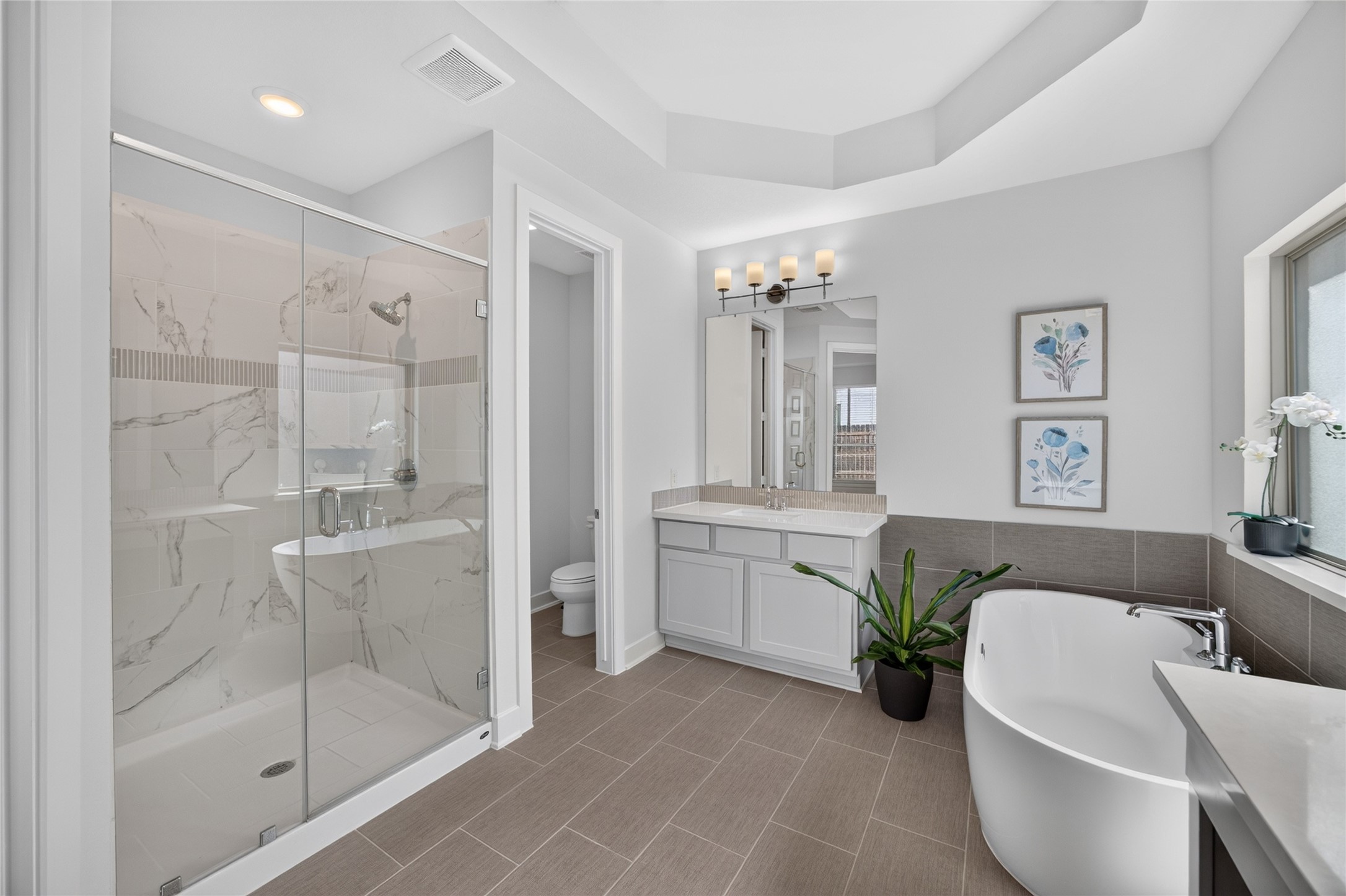 A glass-enclosed shower showcases large-format patterned tile and a coordinating decorative accent band. The adjacent freestanding tub provides a separate bathing option, while the nearby vanity offers storage and counter space. Natural light and neutral finishes give the room a bright, cohesive appearance.