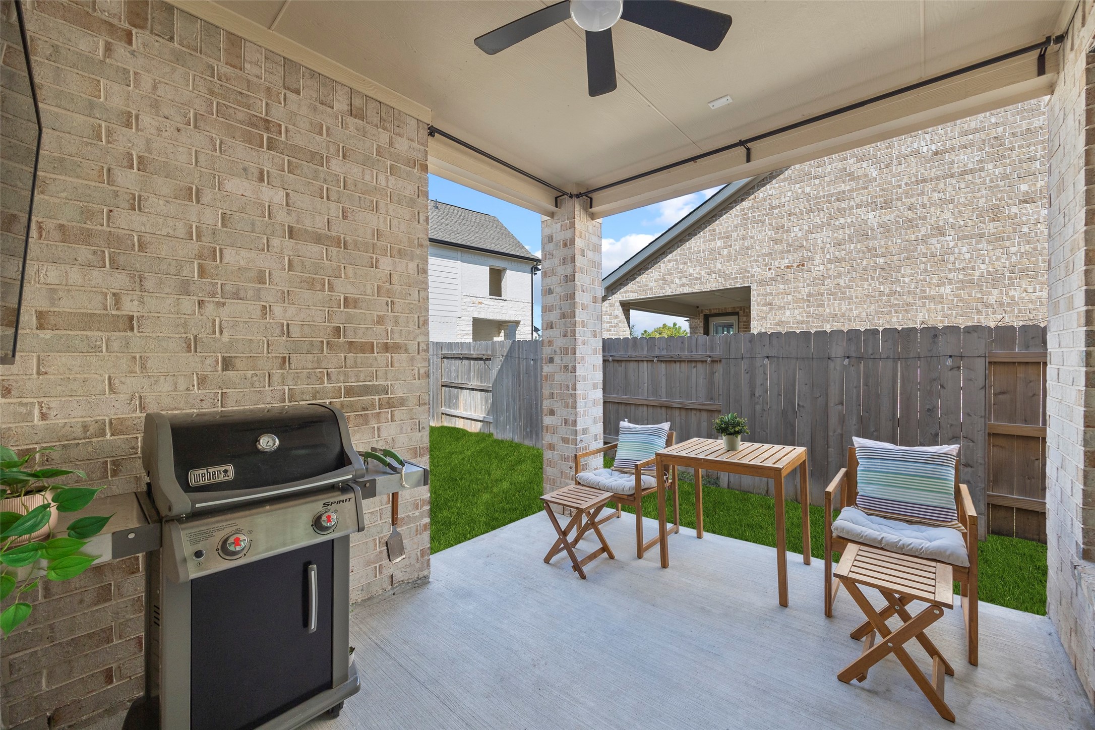 The covered patio offers a sheltered extension of the homes living space, with room for a small dining arrangement and grill setup. Brick walls and columns complement the exterior, while a ceiling fan helps circulate air. The patio opens directly to the lawn for convenient indoor-outdoor gatherings.