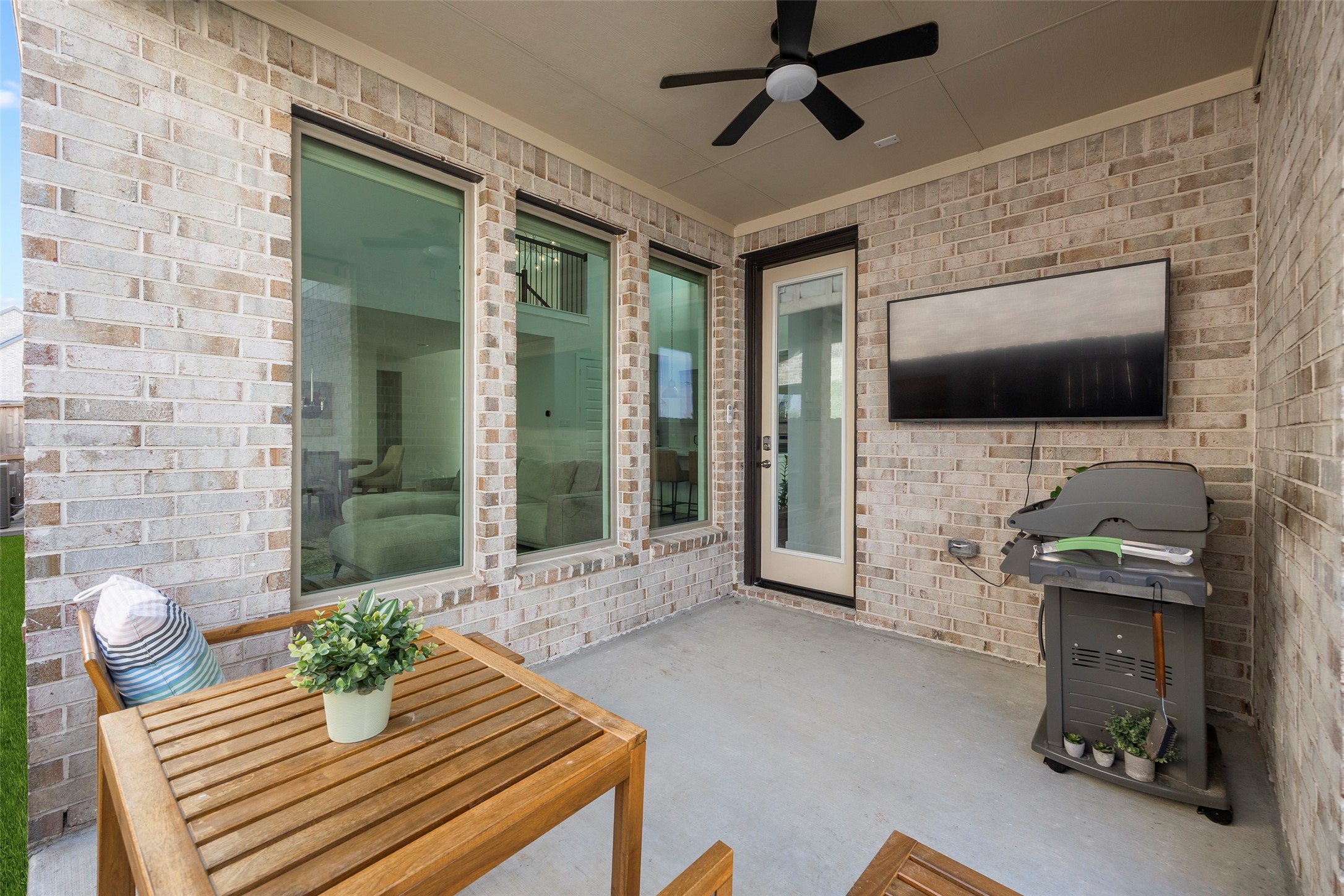 This reverse view of the covered patio shows the rear entry door, tall windows and brick backdrop. The sheltered space is currently arranged for outdoor dining and television viewing, illustrating its potential for relaxed evenings or weekend entertaining. A ceiling fan adds comfort above the seating area.
