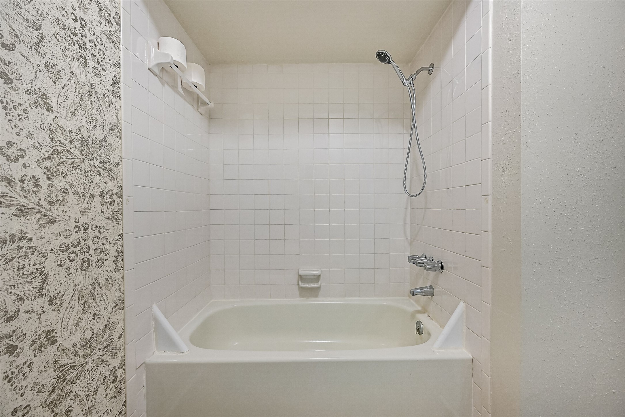 The primary bathroom includes a white tiled tub/shower combination with a handheld showerhead, built-in soap dish & towel bar. The adjoining vanity features a granite countertop & white cabinetry, while the separate bathing area offers a practical layout with everything within easy reach.