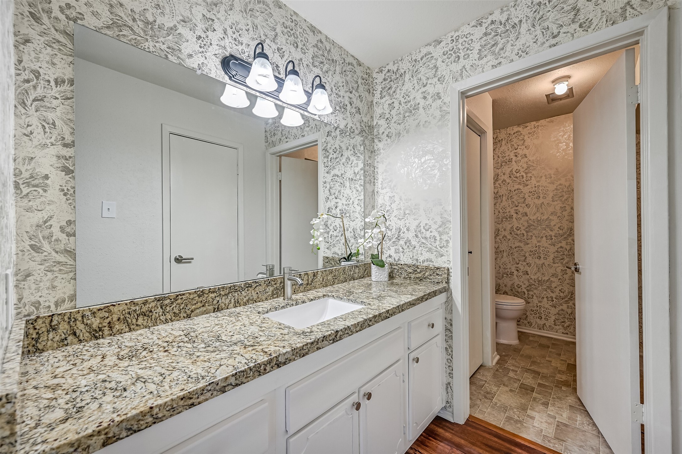 The primary bathroom features a separate vanity area with a granite countertop, undermount sink, updated faucet & contemporary vanity lighting. Decorative floral wallpaper adds character, while a large wall mirror & white cabinetry provide a functional space for getting ready each day.