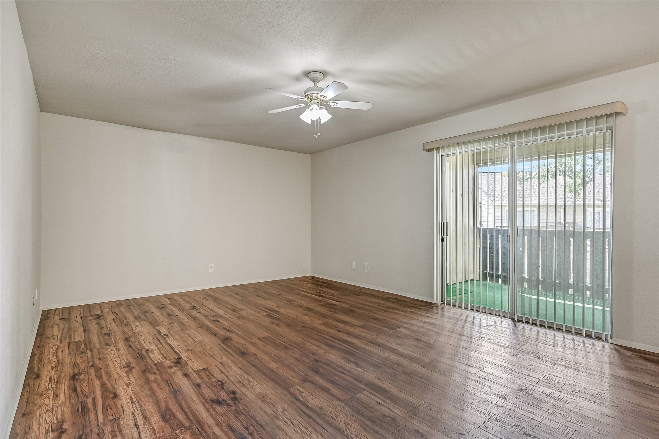 Another view of the spacious living room highlights the sliding glass doors opening to the oversized covered balcony. Wood-look flooring extends throughout the room, while a ceiling fan with lighting adds comfort. The generous wall space allows for a variety of furniture arrangements.