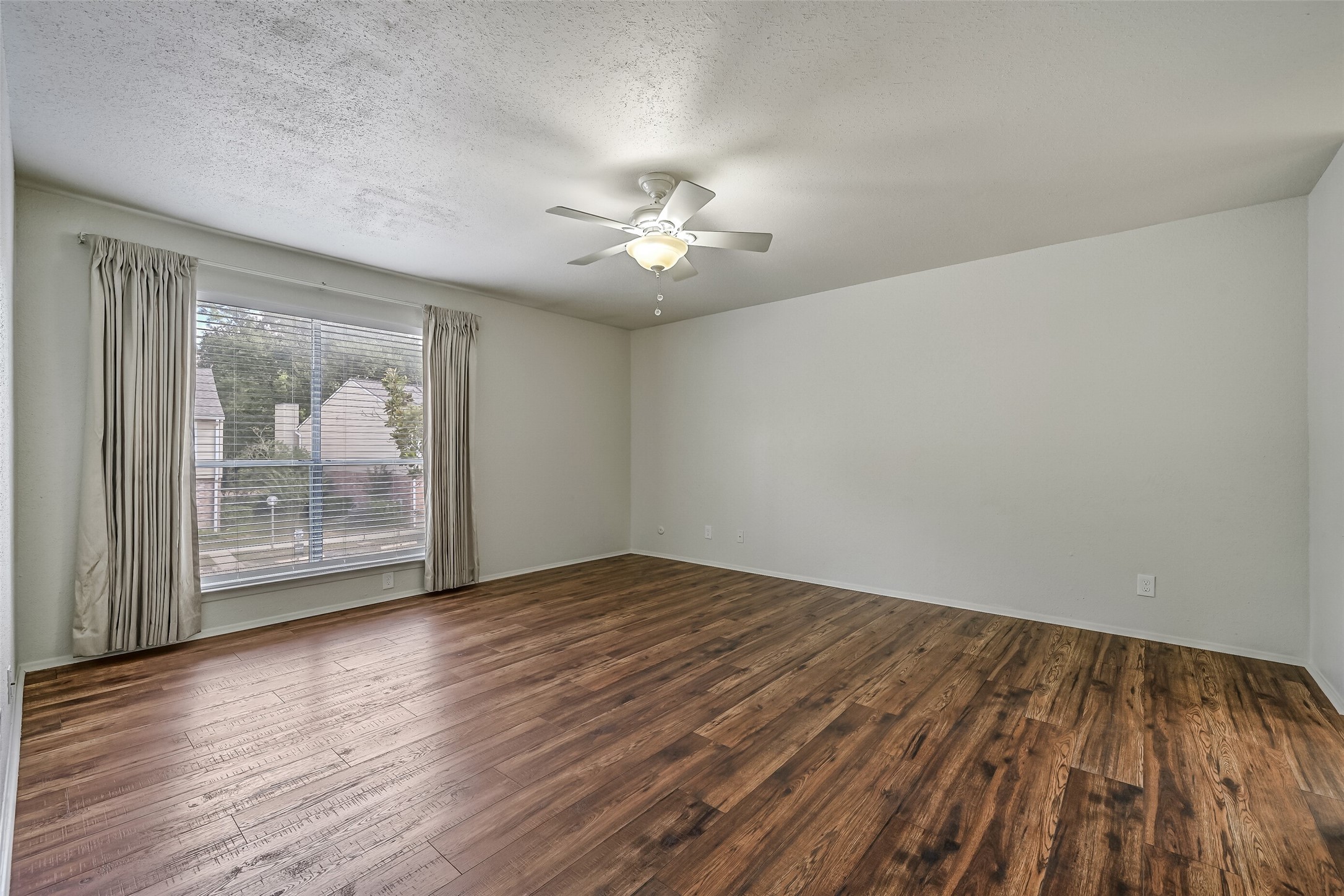 A large window with blinds & curtains brings natural light into the primary bedroom, while the wood-look flooring adds warmth to the neutral interior. With plenty of open wall space for furniture placement, this generously sized room offers flexibility for creating your ideal bedroom retreat.