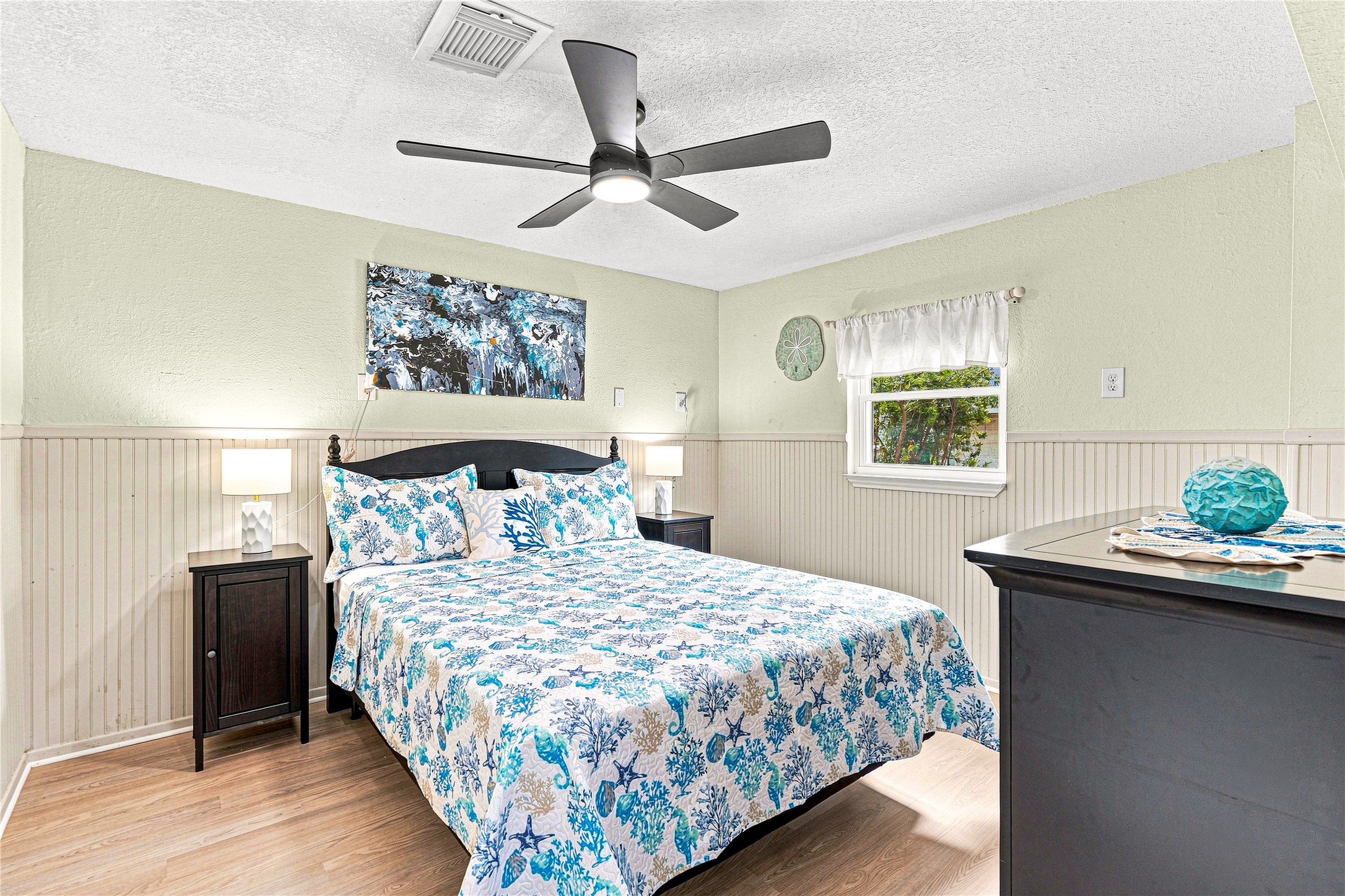 Cozy bedroom with a modern ceiling fan, featuring a comfortable bed adorned with a vibrant blue and white quilt. Natural light streams through a window, complementing the soft, neutral wall tones and wainscoting.