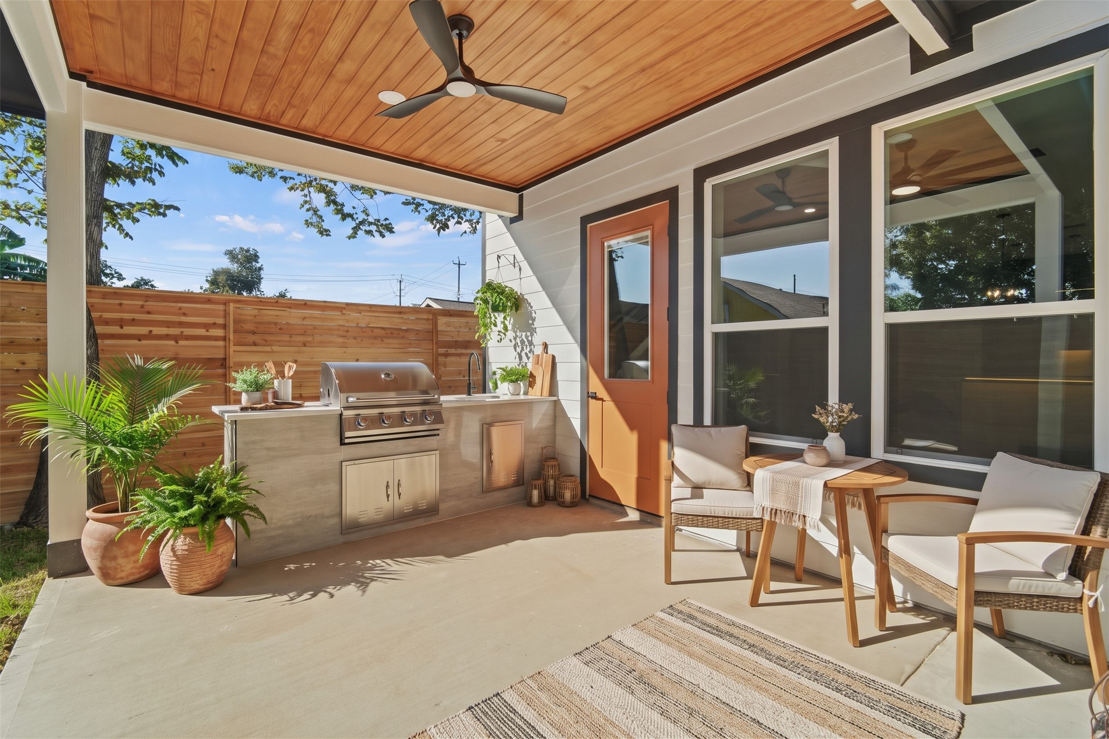 Virtually Staged: This covered back patio features a built-in outdoor kitchen island equipped with stainless steel storage drawers, a sink, and prep counter space. A natural wood ceiling with a modern fan anchors the sheltered space.
