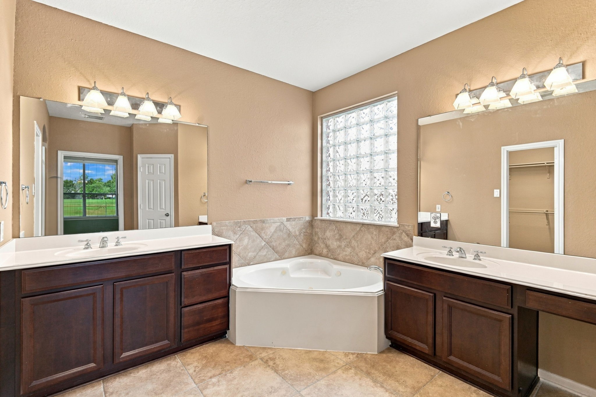 This spacious primary ensuite bath features dual separate vanities with rich wood cabinetry and ample counter space. A luxurious corner soaking tub serves as the centerpiece, flanked by a privacy glass block window.