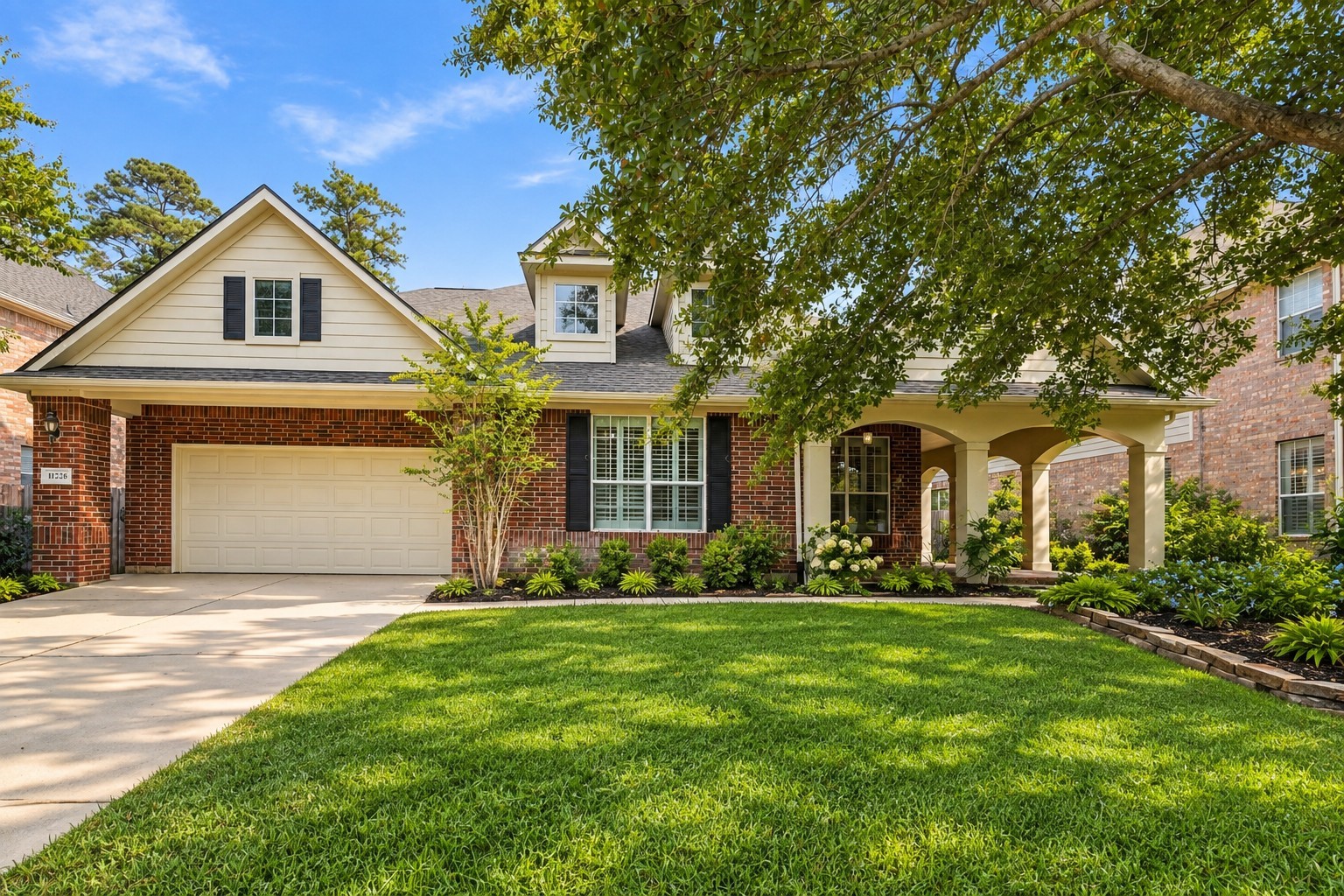 Practical features like a large driveway and generous garage space are paired with a cozy covered porch nestled among mature, leafy trees and vibrant gardens.